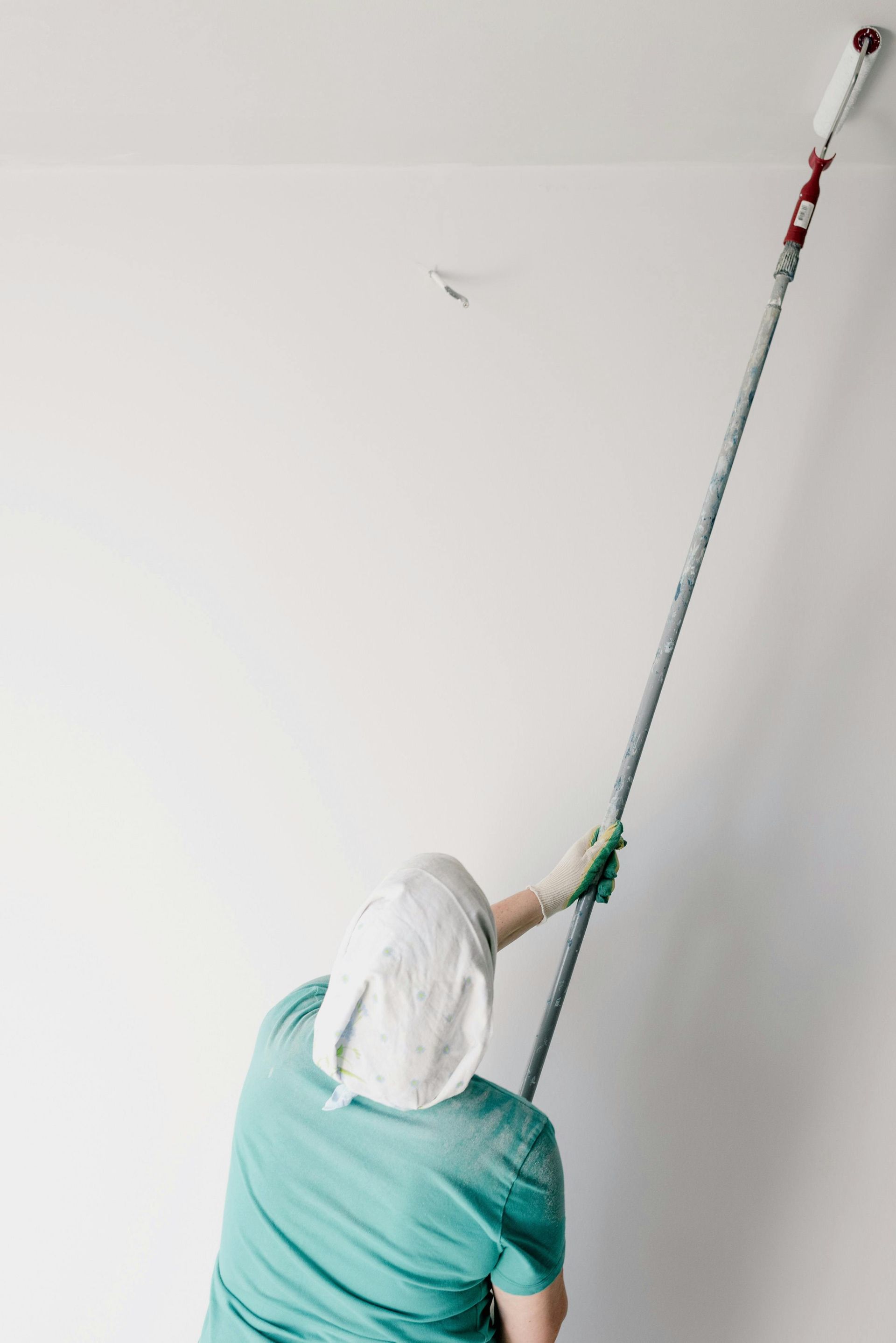 A person wearing a head covering and teal shirt uses a long extension pole with a paint roller to paint a white ceiling.