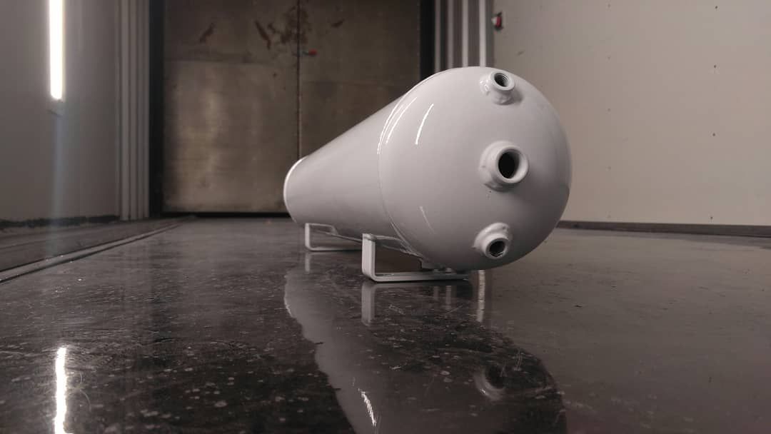 White cylindrical air tank with ports, on a metal stand, on a glossy floor, in a light-filled room.