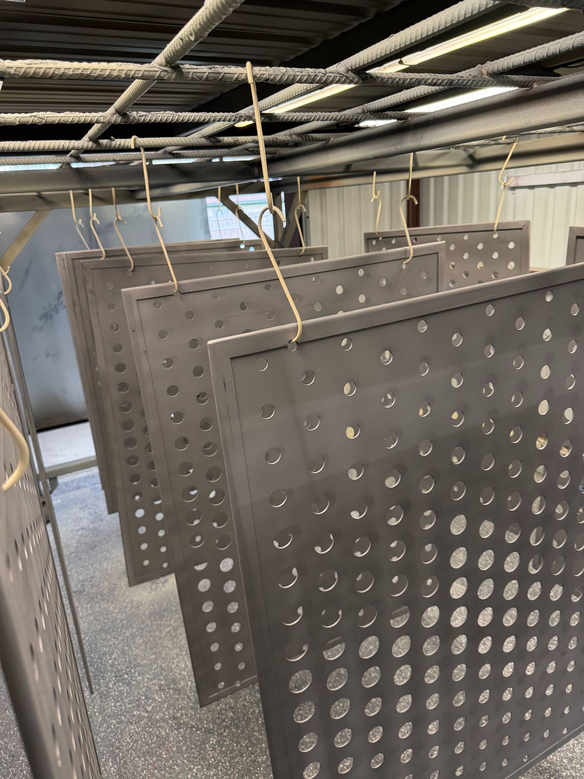 Several dark, perforated metal panels hang from wires on a metal rack in an industrial workshop.