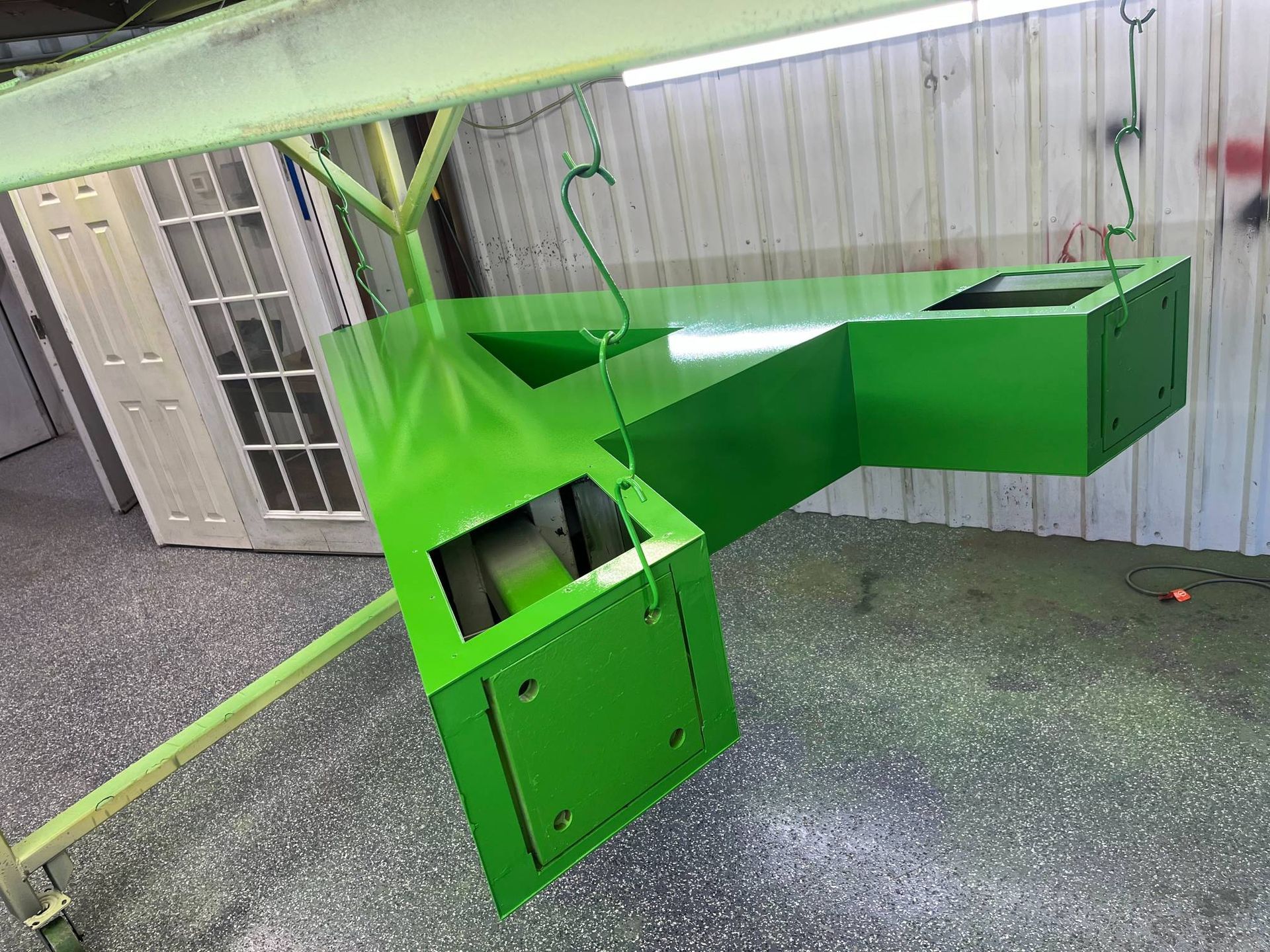 A bright green, L-shaped industrial metal component hangs from hooks in a workshop.