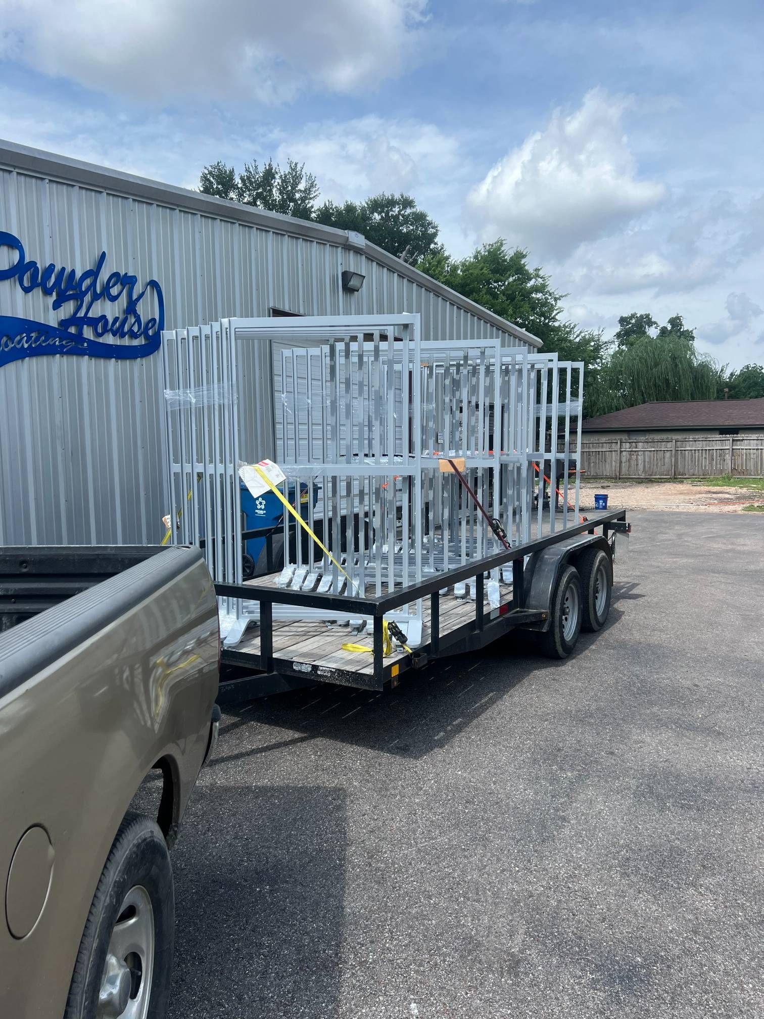 A utility trailer hitched to a truck, loaded with white metal frames, parked outside a building with a blue sign.