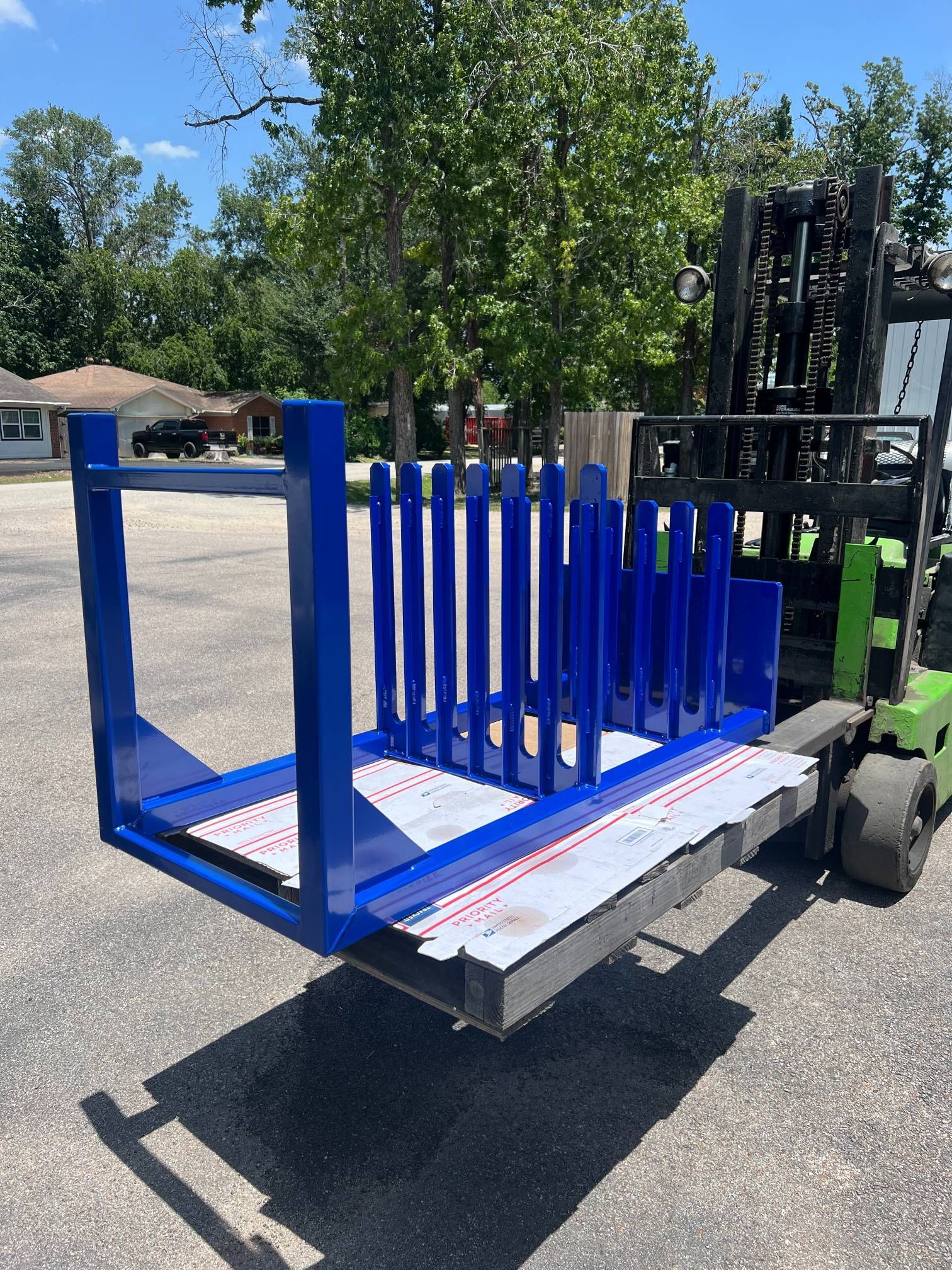 A blue steel A-frame rack for material transport sits on a pallet being held by a forklift in an outdoor parking lot.