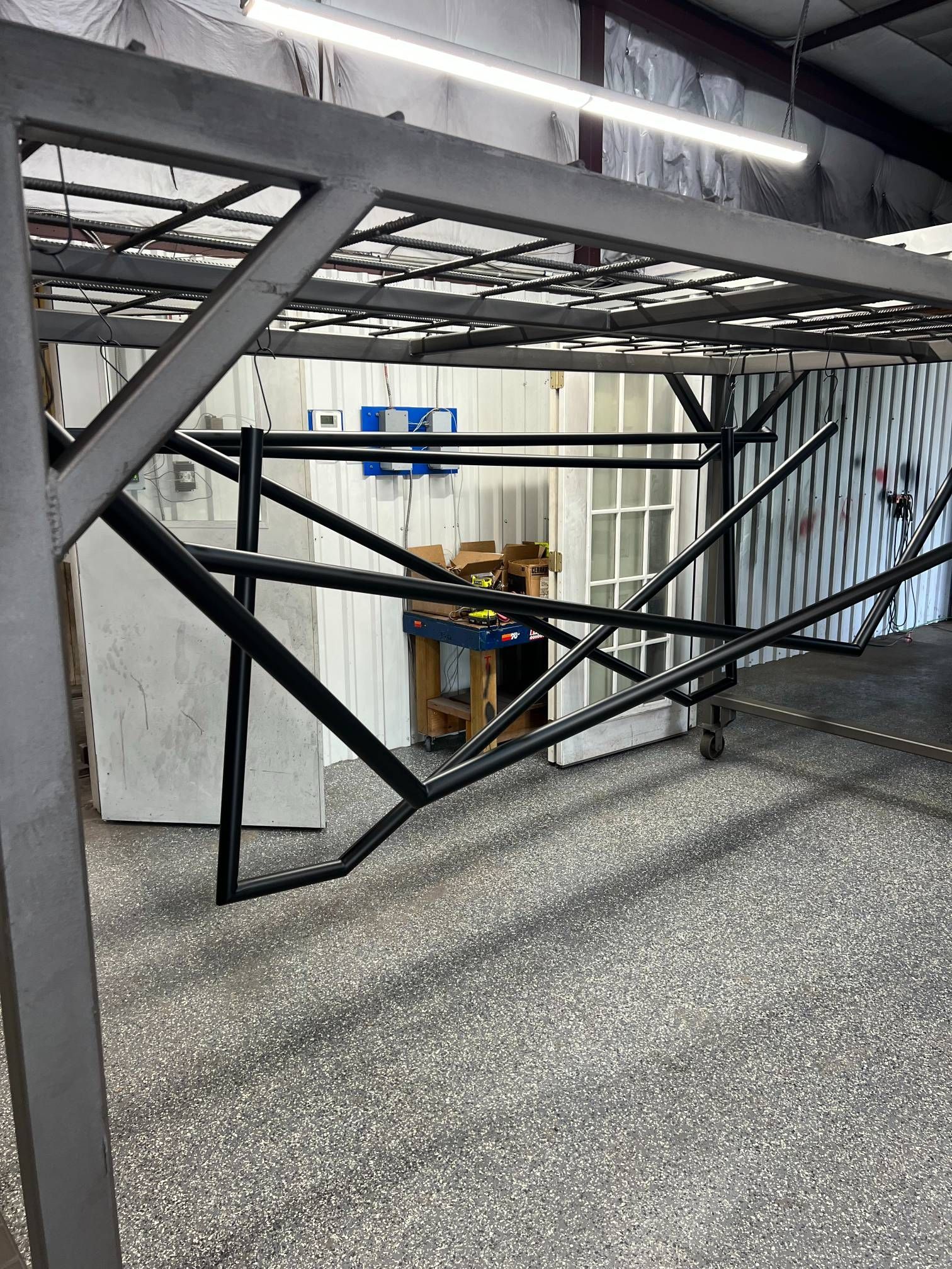 A black metal frame structure hangs from an overhead rack in a workshop, with tools and a doorway in the background.