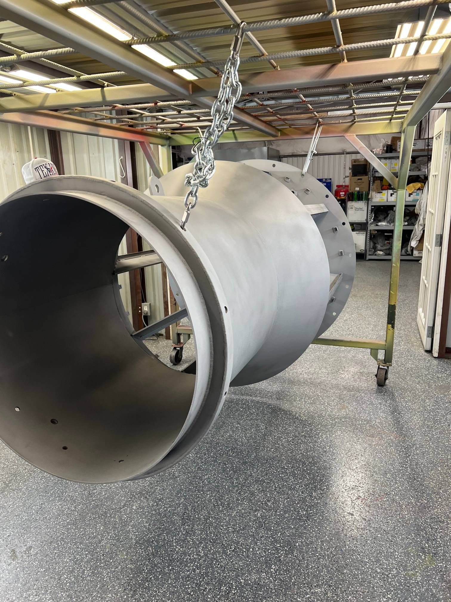 A large, grey, industrial metal pipe component hangs from chains in a workshop with a speckled floor.
