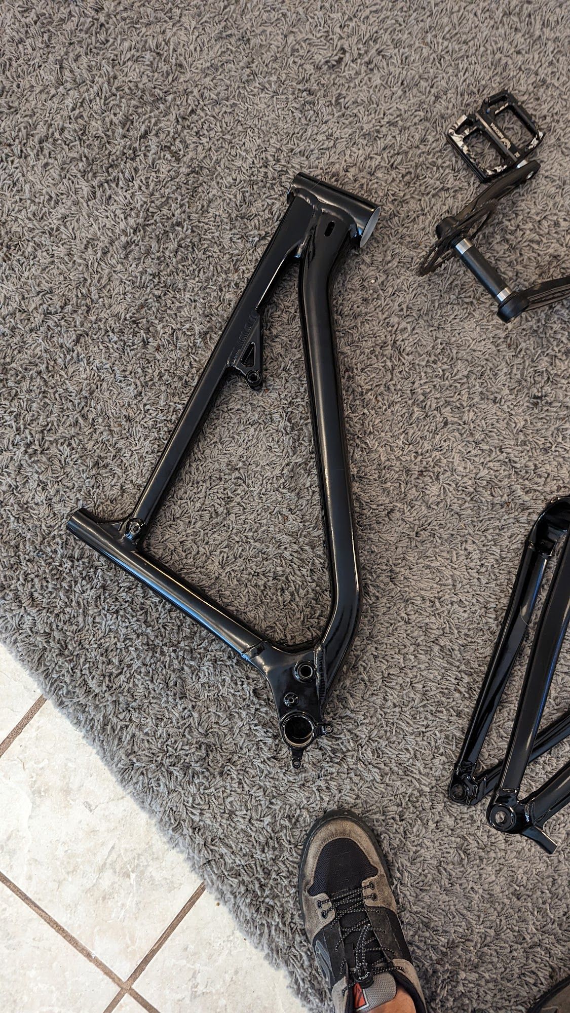 Black bicycle frame and pedals on a gray carpet, next to a person's foot in a shoe.