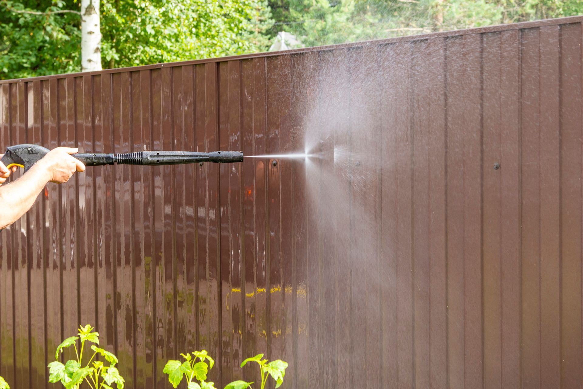 A man is using a high pressure washer to clean a wooden fence.