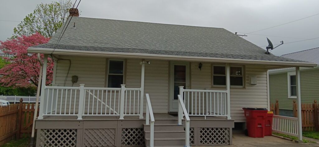 A small, white house with a porch and a red trash bin. There is a tree on the left side.