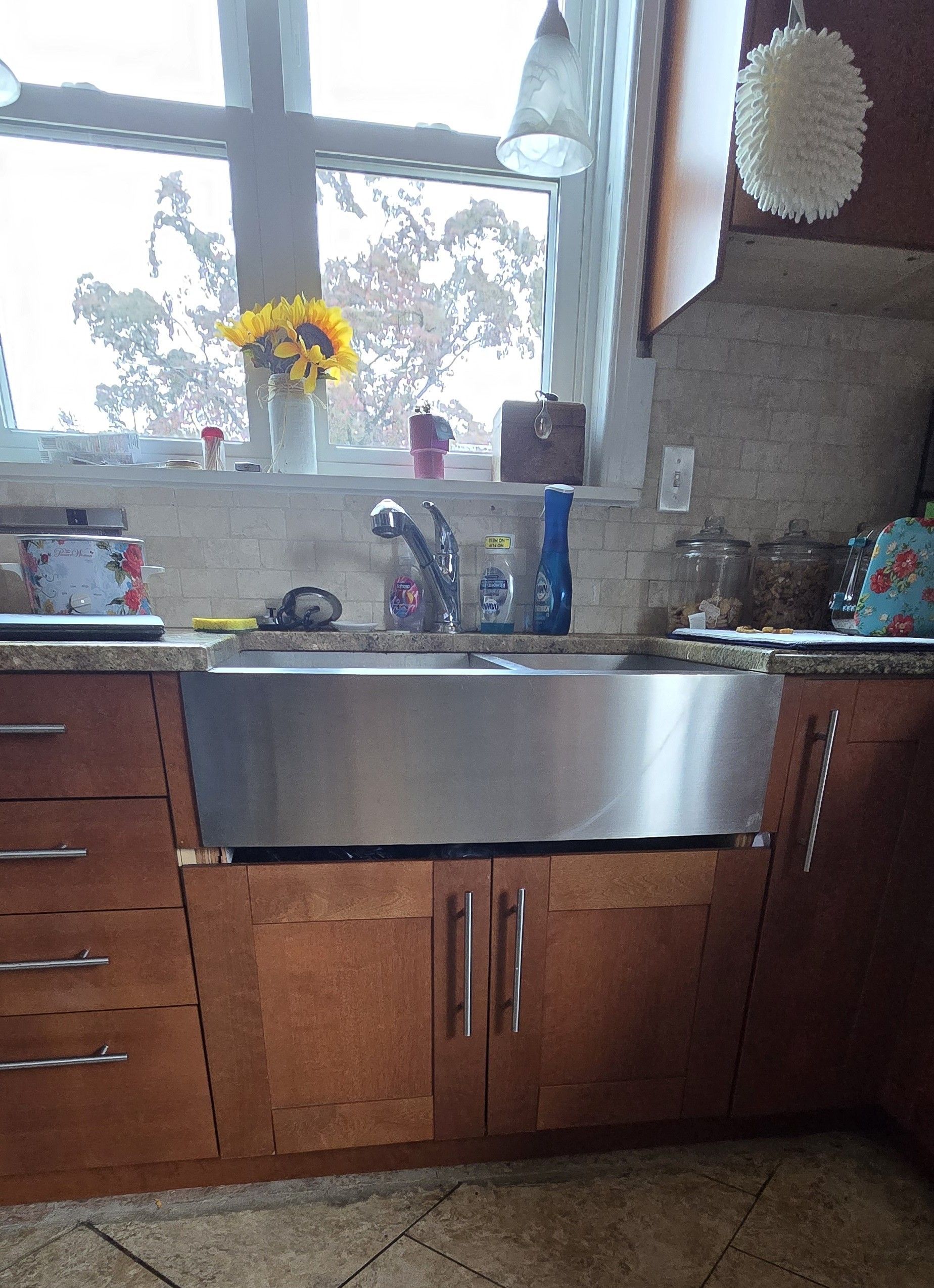 Stainless steel farmhouse sink in a kitchen with wooden cabinets, a window with flowers.