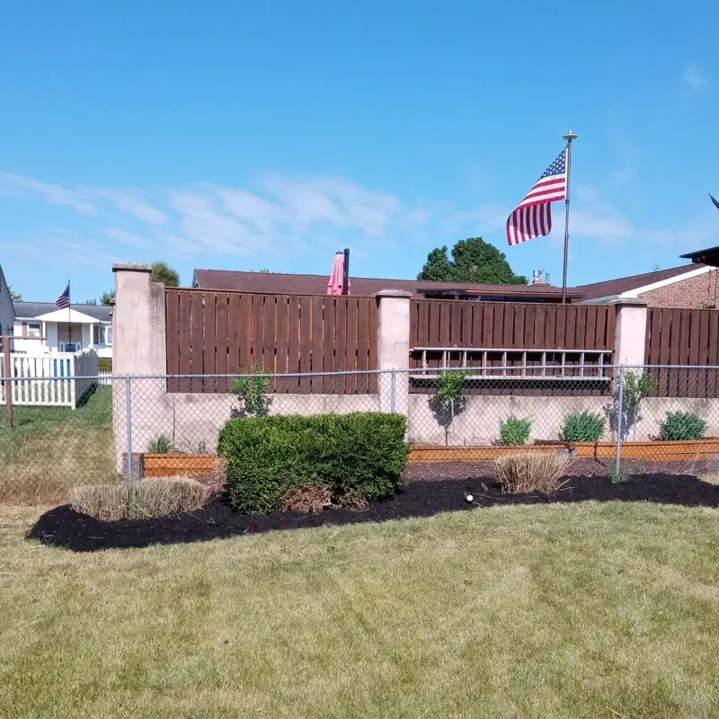 Backyard with fence, landscaping, American flag on pole, blue sky.