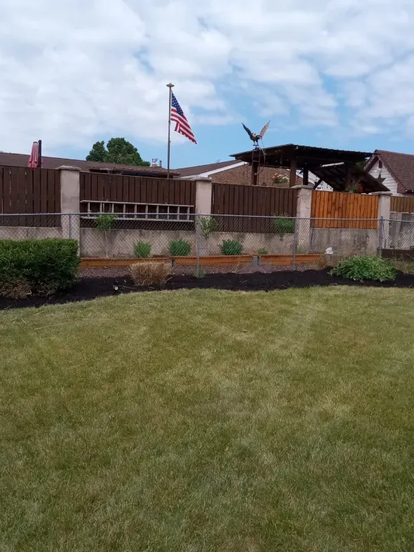A yard with an American flag, fence, dark mulch, and green grass on a cloudy day.