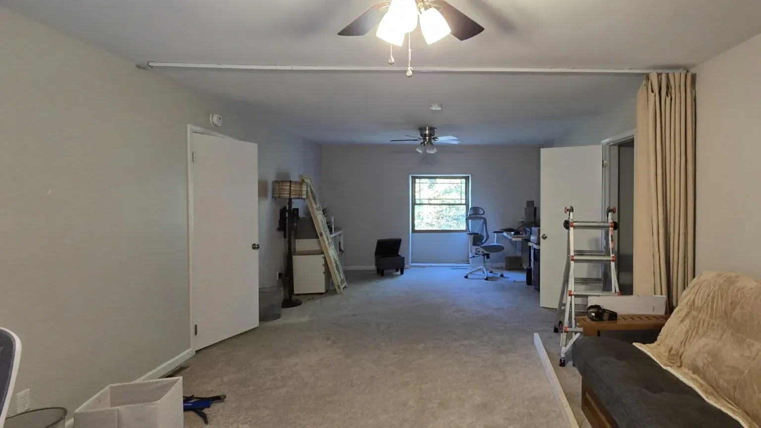 Spacious room with gray walls, carpet, ceiling fans. Curtain divides the space. Cluttered with furniture and objects.