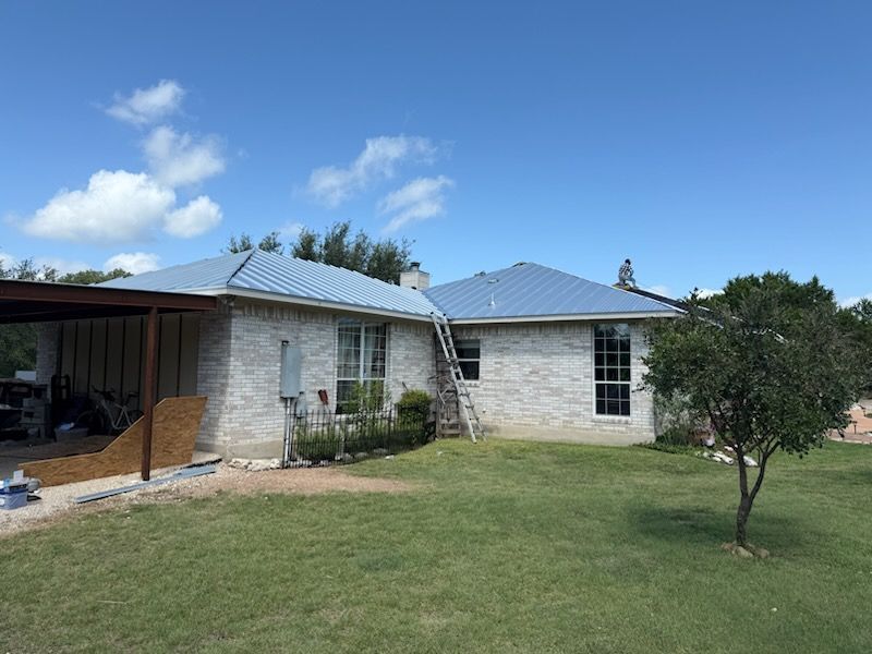 Blue metal roof on a light brick house with green grass and a blue sky. A ladder is propped against the roof.