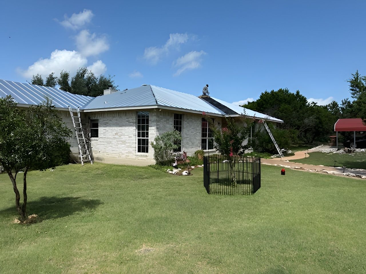 A one-story brick home with a blue metal roof, ladder, and black fence on a green lawn.