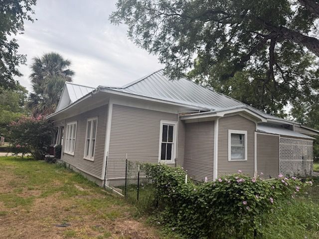Tan house with metal roof, windows, and overgrown bushes in front.