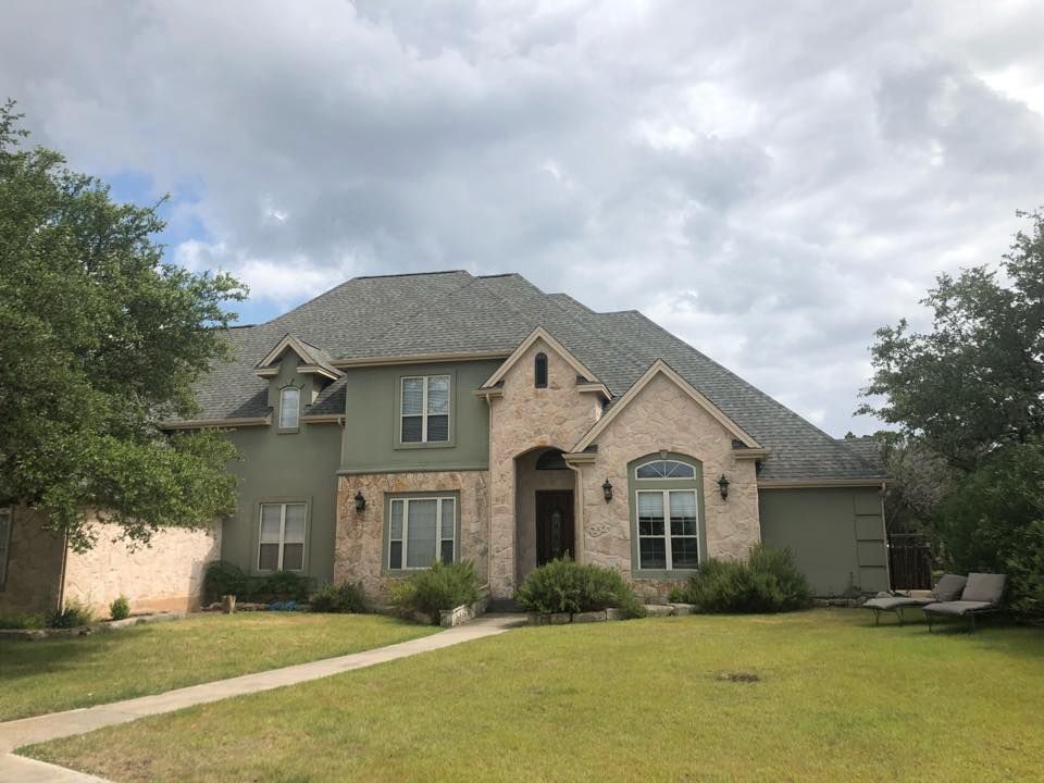 Two-story house with green stucco and brick facade under a cloudy sky, with lawn and trees.