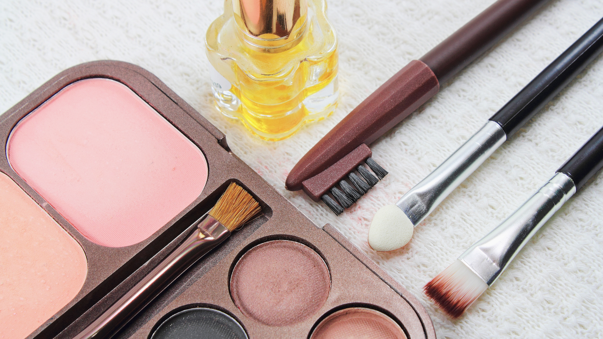 A palette of makeup , brushes , and a bottle of perfume on a table.