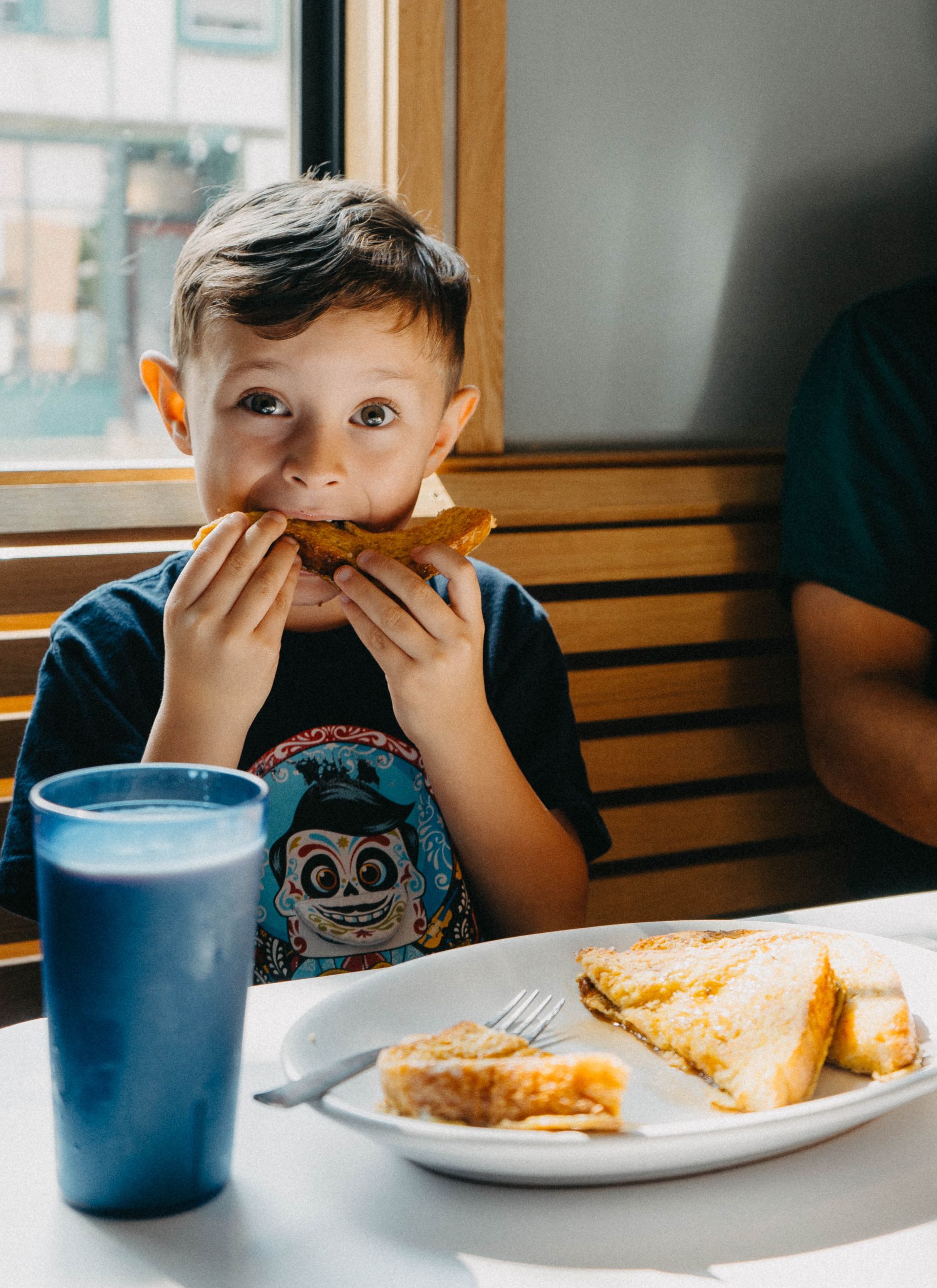 A young boy is sitting at a table eating a sandwich and drinking milk