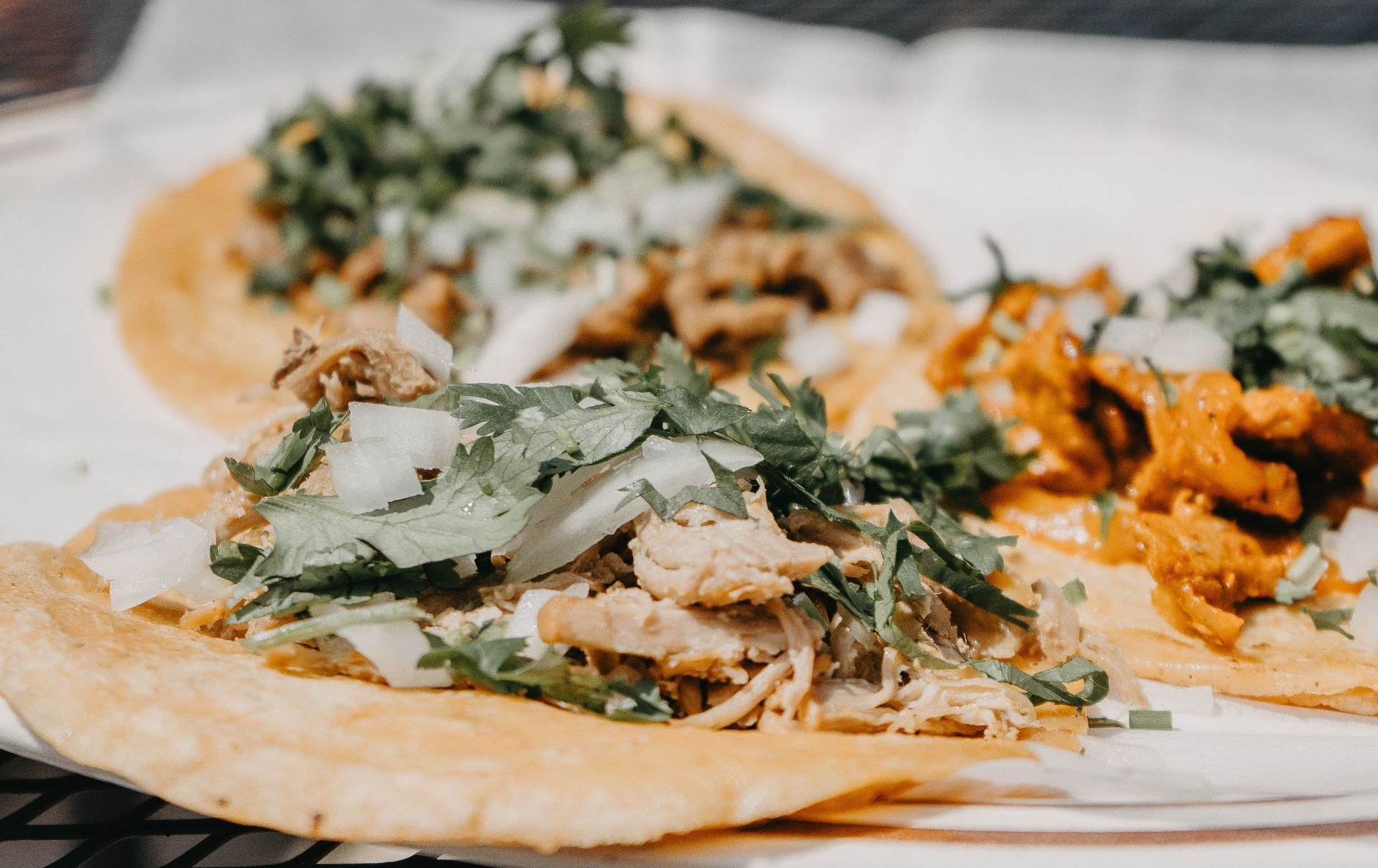 Three tacos on a white plate, topped with cilantro, onions, and different meats.