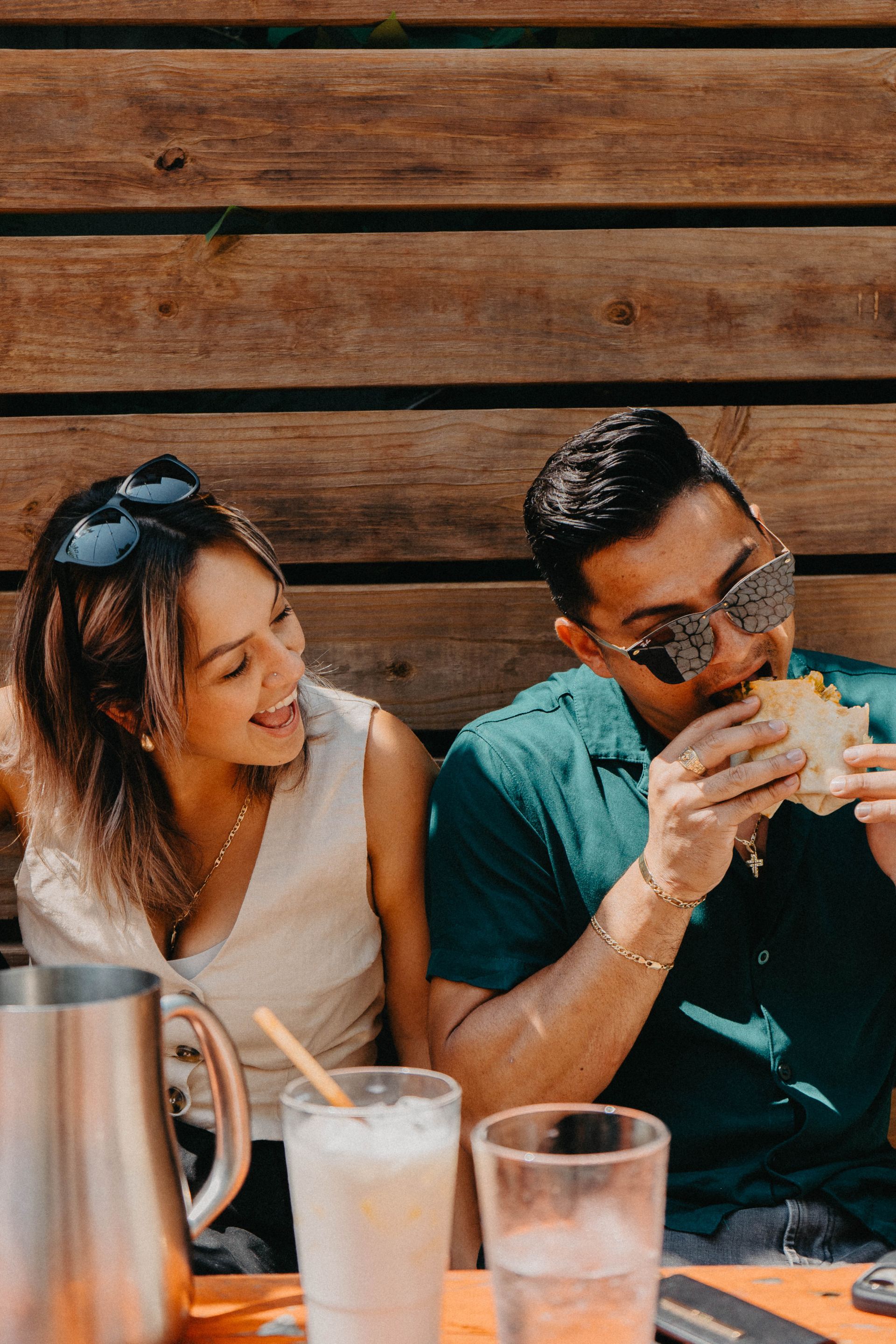 Woman smiles at man eating a taco outdoors. They are sitting at a wooden table.