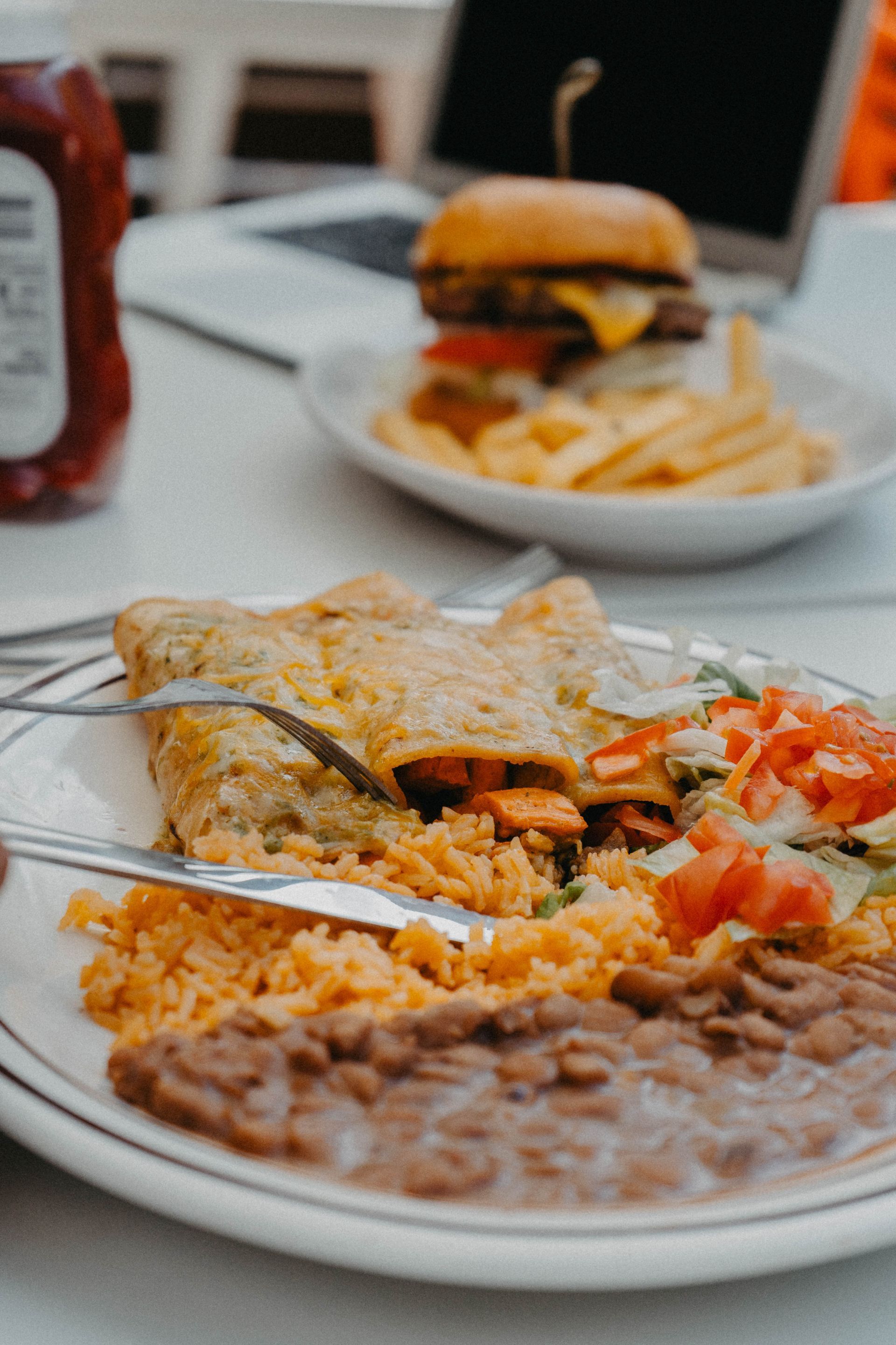 Plate with enchilada, rice, beans, and salsa. Burger and fries in the background.
