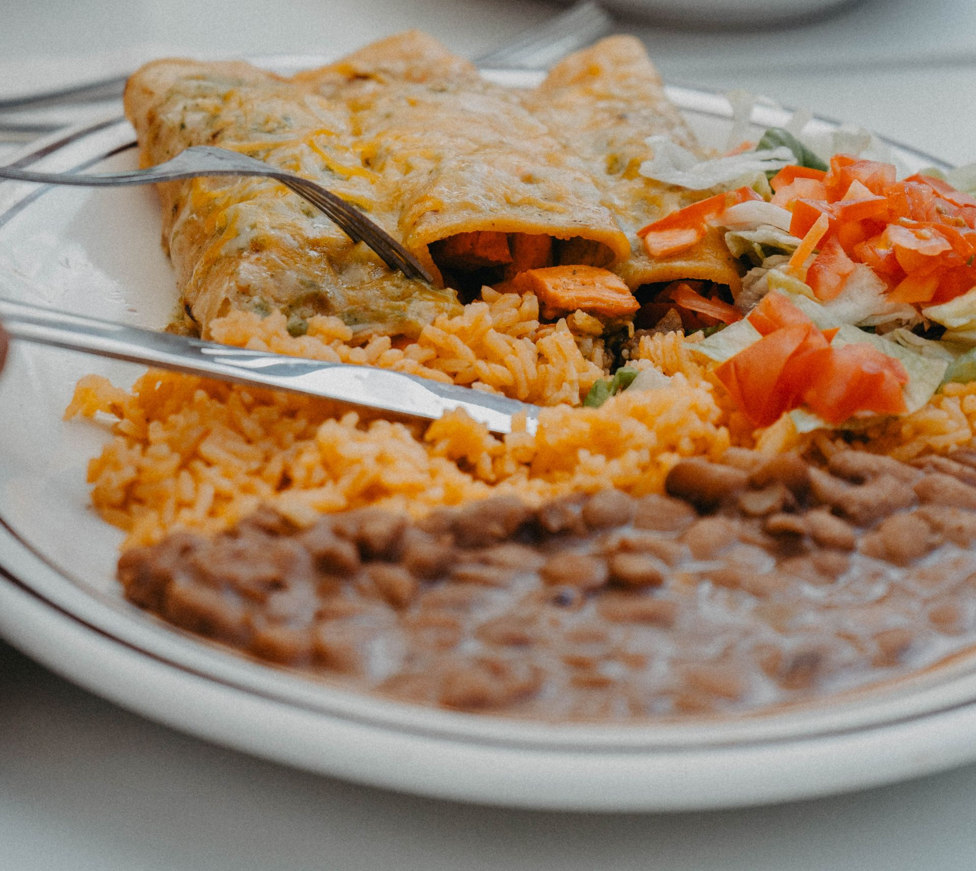 Plate of Mexican food: enchiladas, rice, beans, lettuce, tomatoes. Fork and knife cutting into the enchilada.