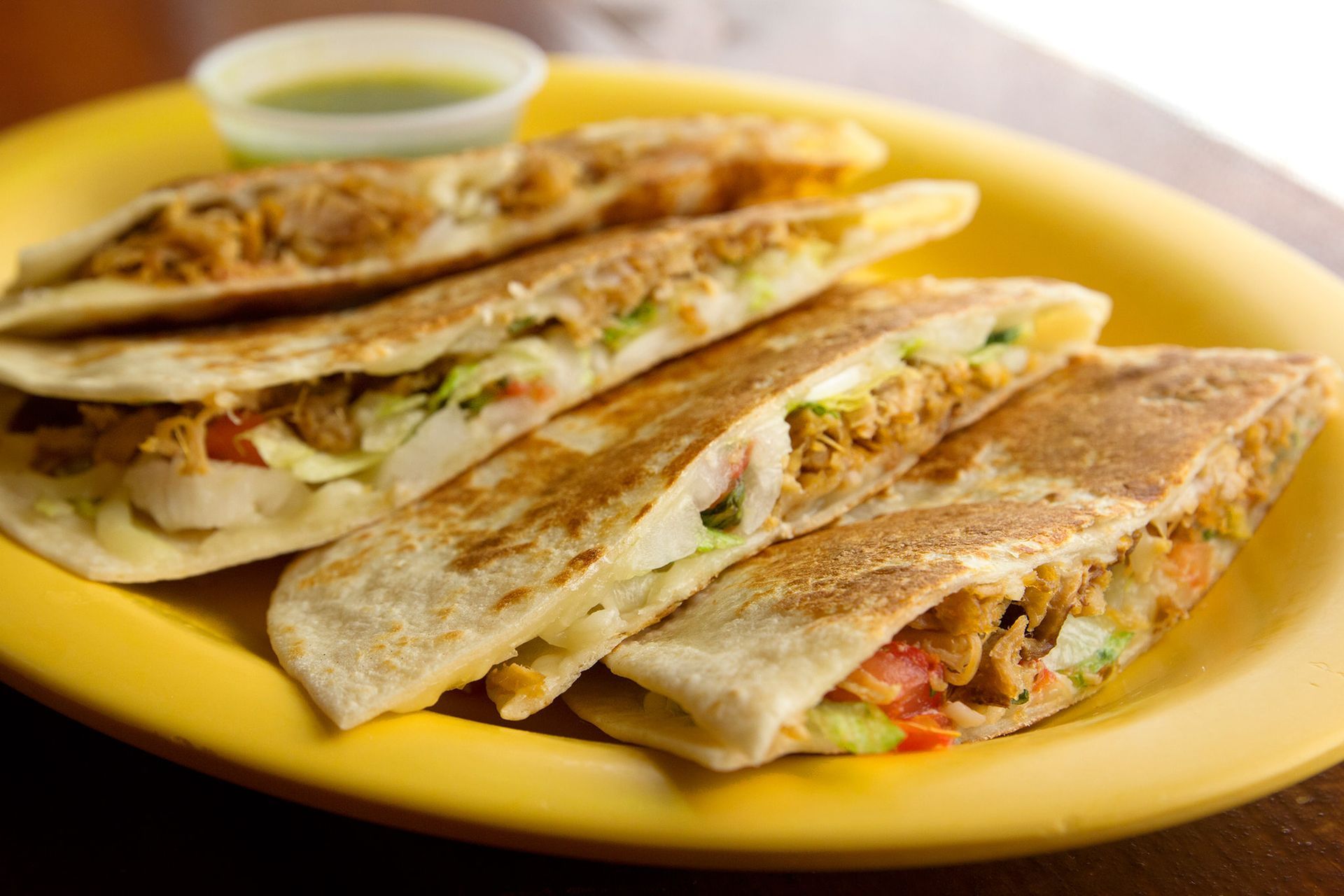 Sliced quesadillas on a yellow plate with a small side of green sauce.