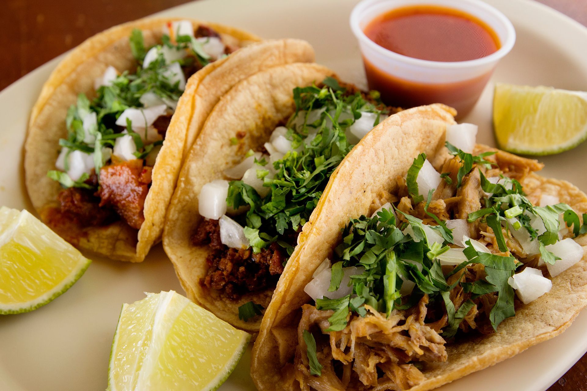 Three tacos on a plate with salsa and lime wedges; filled with meat, onions, and cilantro.