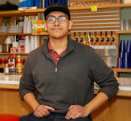 Man in glasses, baseball cap, and sweater sits at a counter in a restaurant setting.