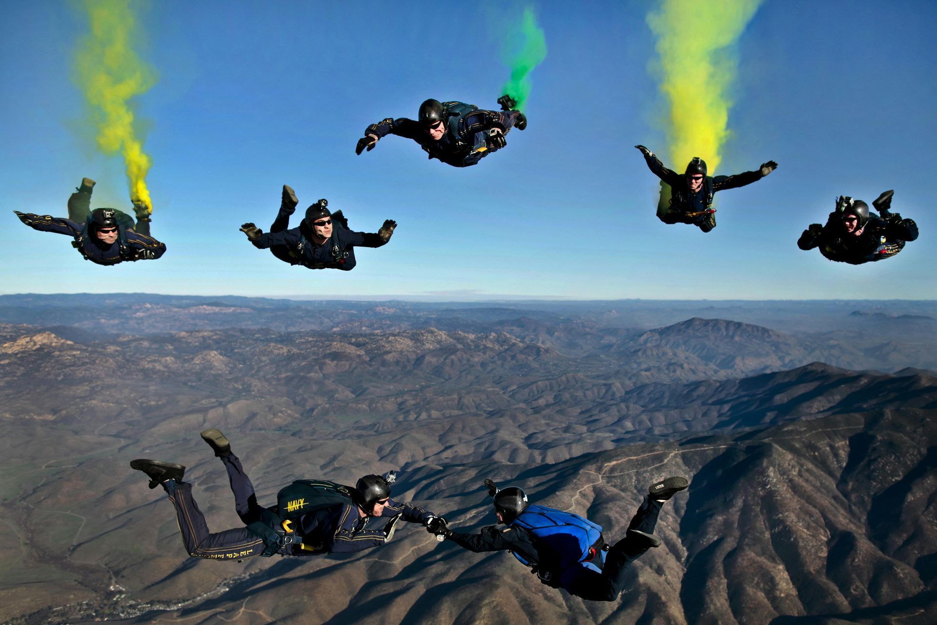 Sky divers in dark suits and helmets, with colored smoke trails, soaring above a mountainous landscape.