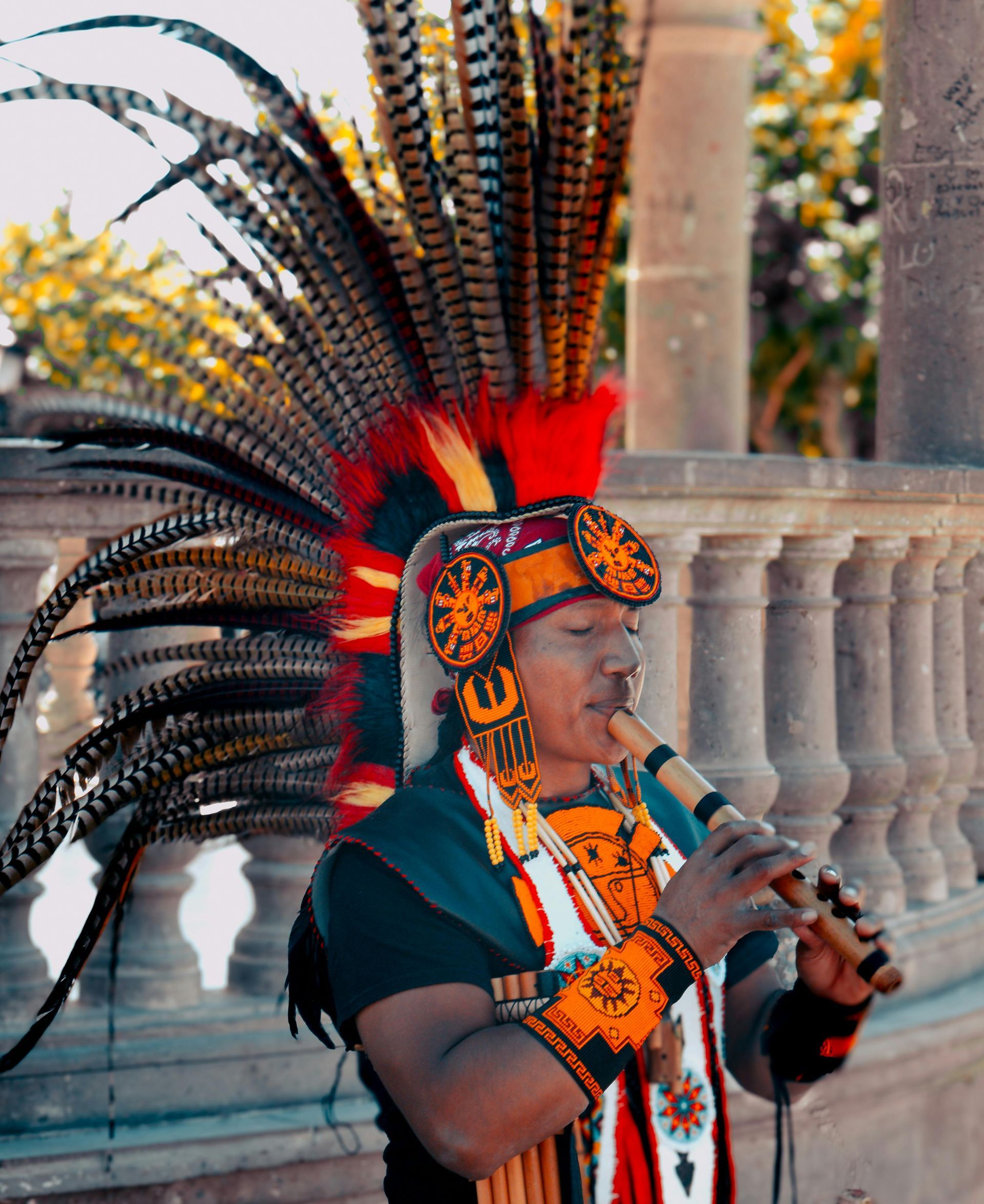 Man playing a flute, wearing elaborate feathered headdress and traditional attire outdoors.