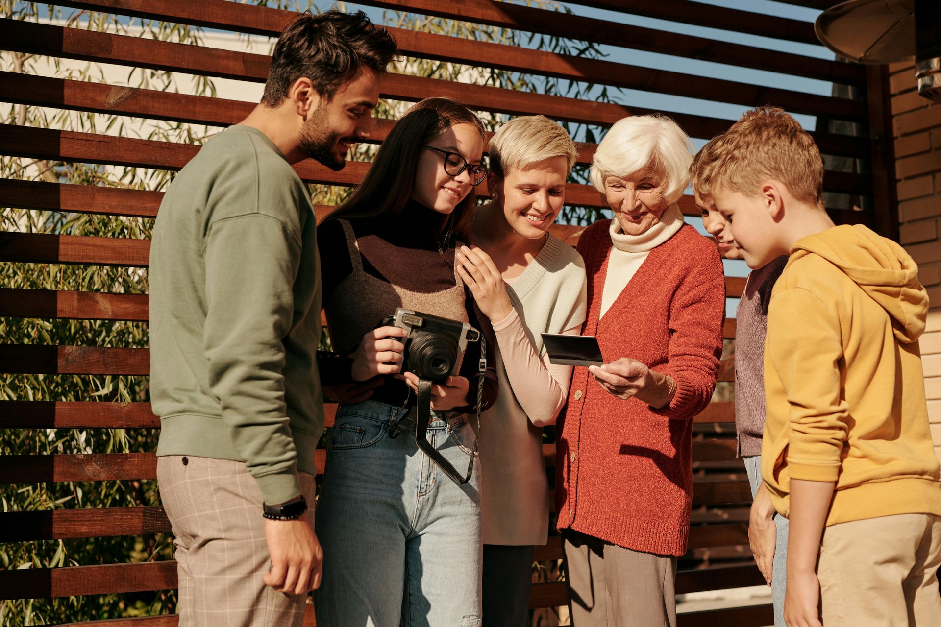 Family looking at a polaroid photo outdoors near a wooden fence. Smiling, they are in casual clothes on a sunny day.