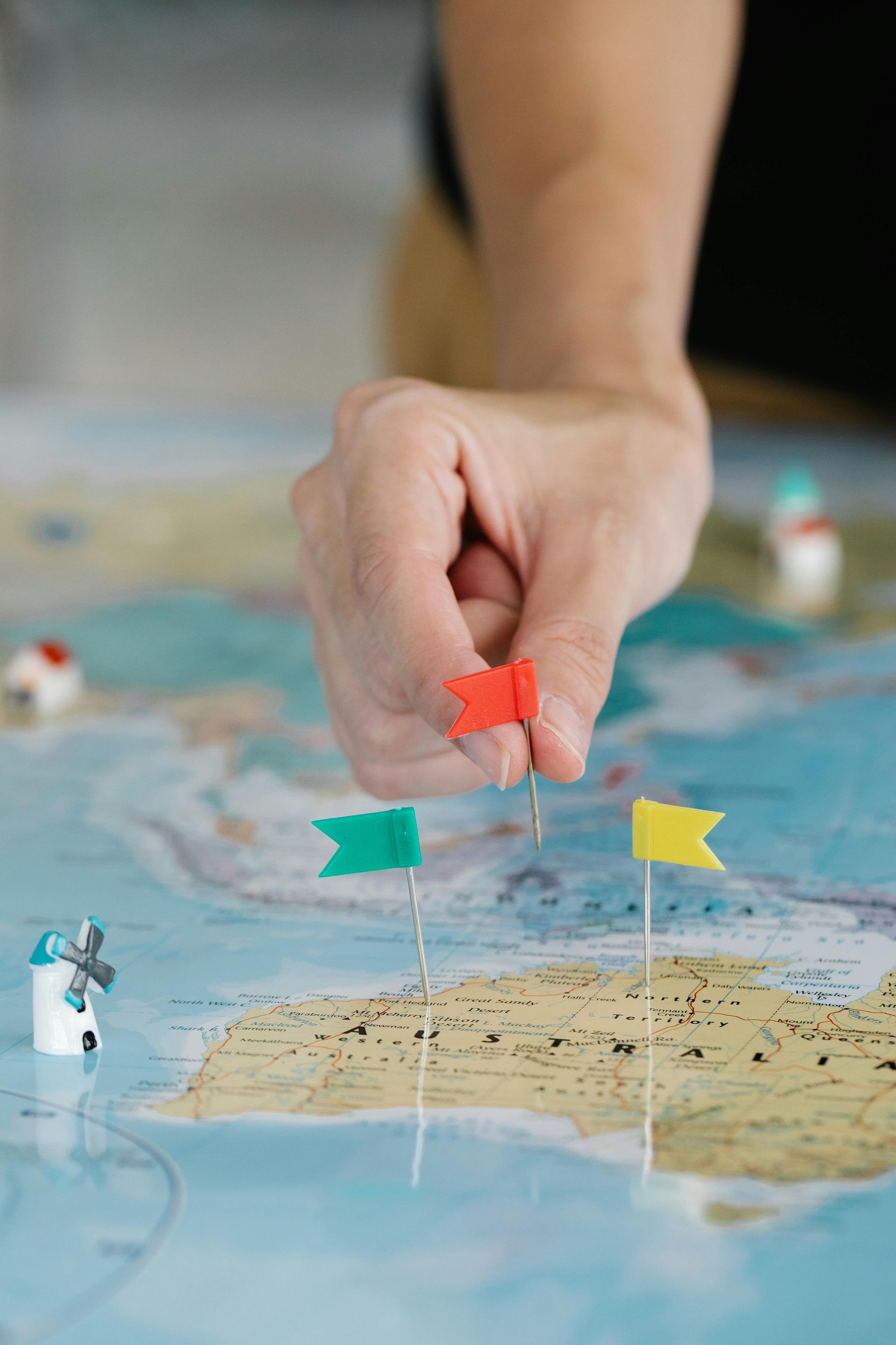Hand placing a red flag on a world map, with other colored flags and miniature buildings.