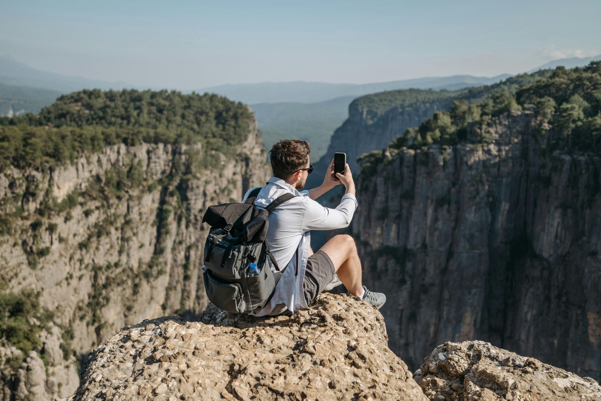 Man on a cliff edge taking a photo of a canyon with a phone. He's wearing a backpack.