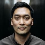 Man with dark hair and a neutral expression, posing in front of a dark backdrop.