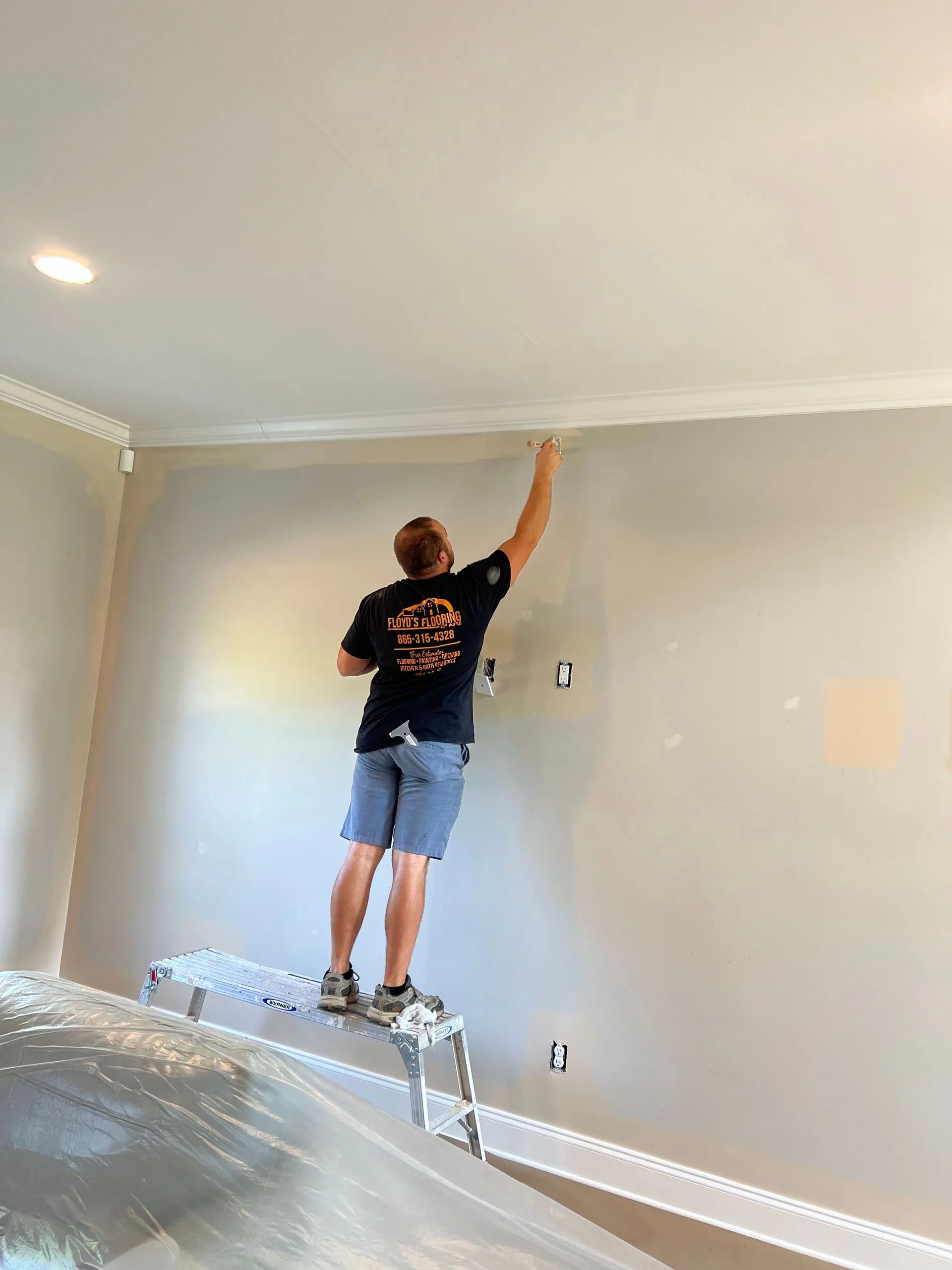 A person standing on a step ladder, painting the edge of a wall near the ceiling in a room.