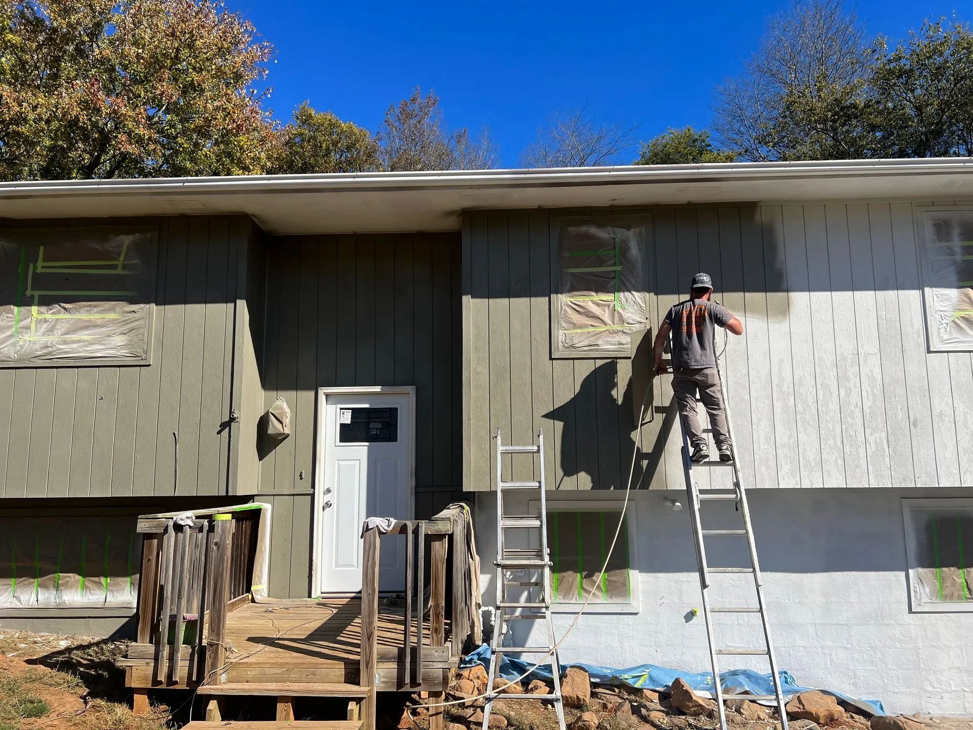 Man painting exterior of a house; one section is green, another white. Ladders used.