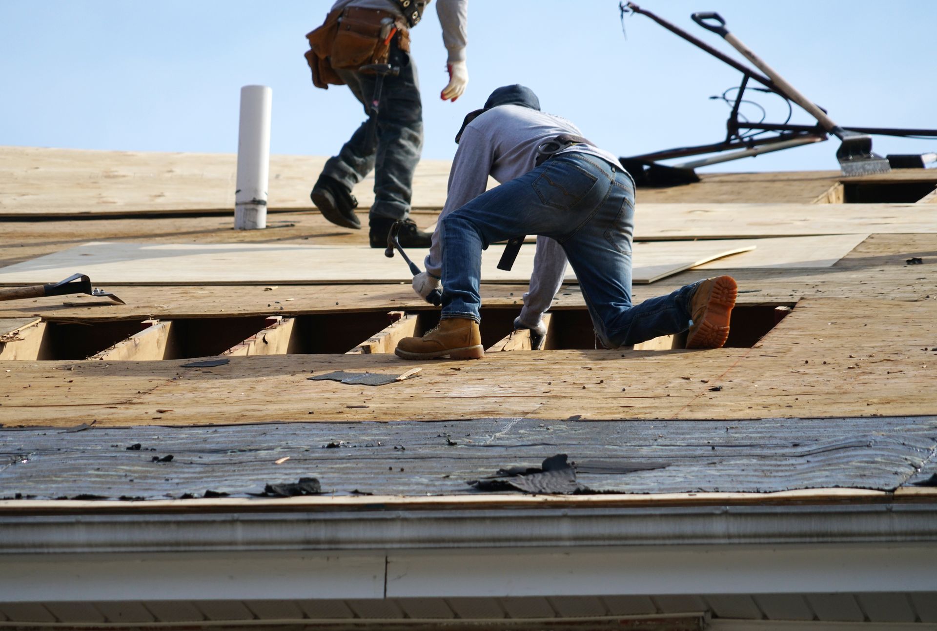 Roofing crew repairing a residential roof deck using tools and plywood under clear sky conditions. Roofing crew repairing a residential roof deck using tools and plywood under clear sky conditions.