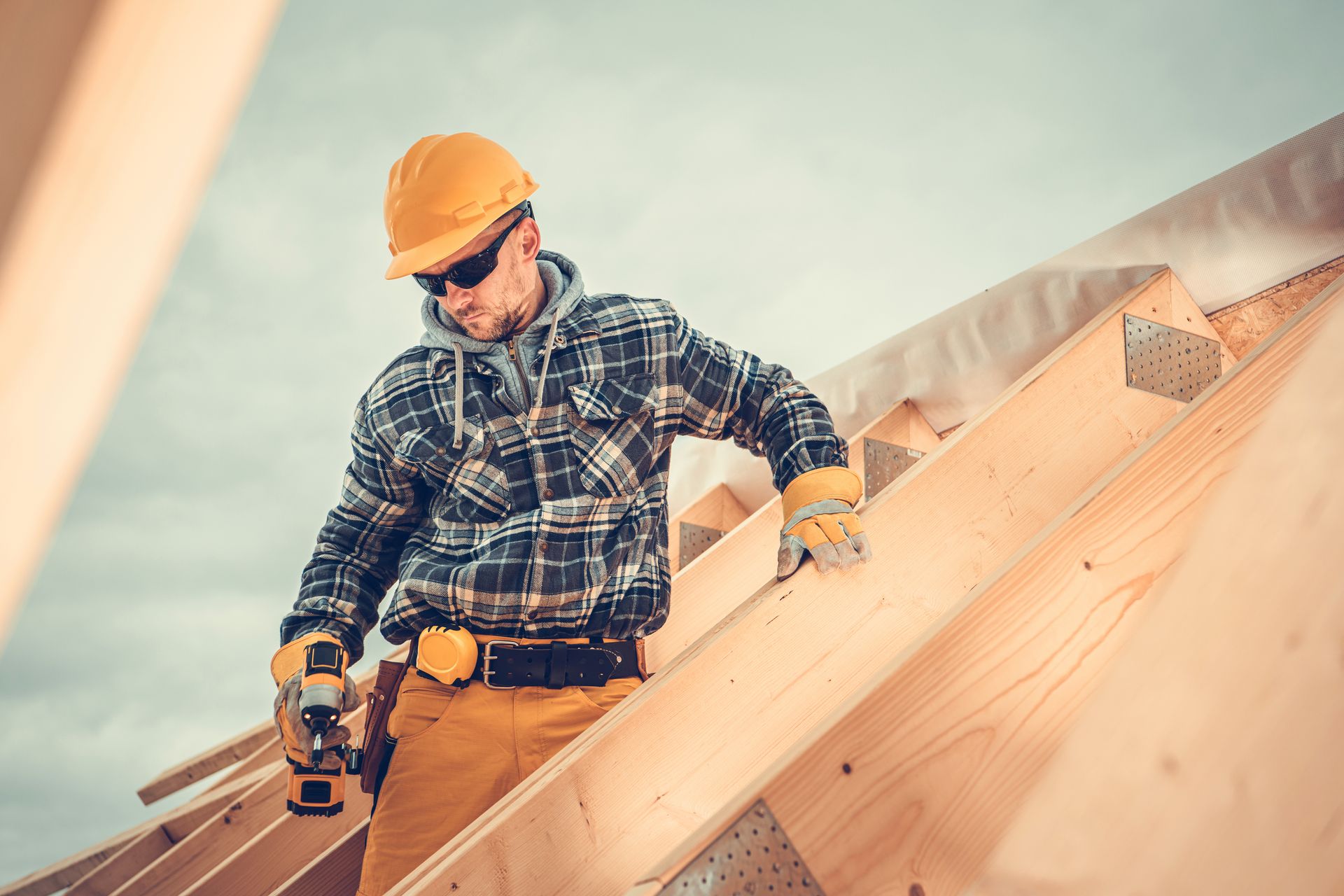 Construction worker wearing a hard hat uses a power drill while installing wooden roof framing.