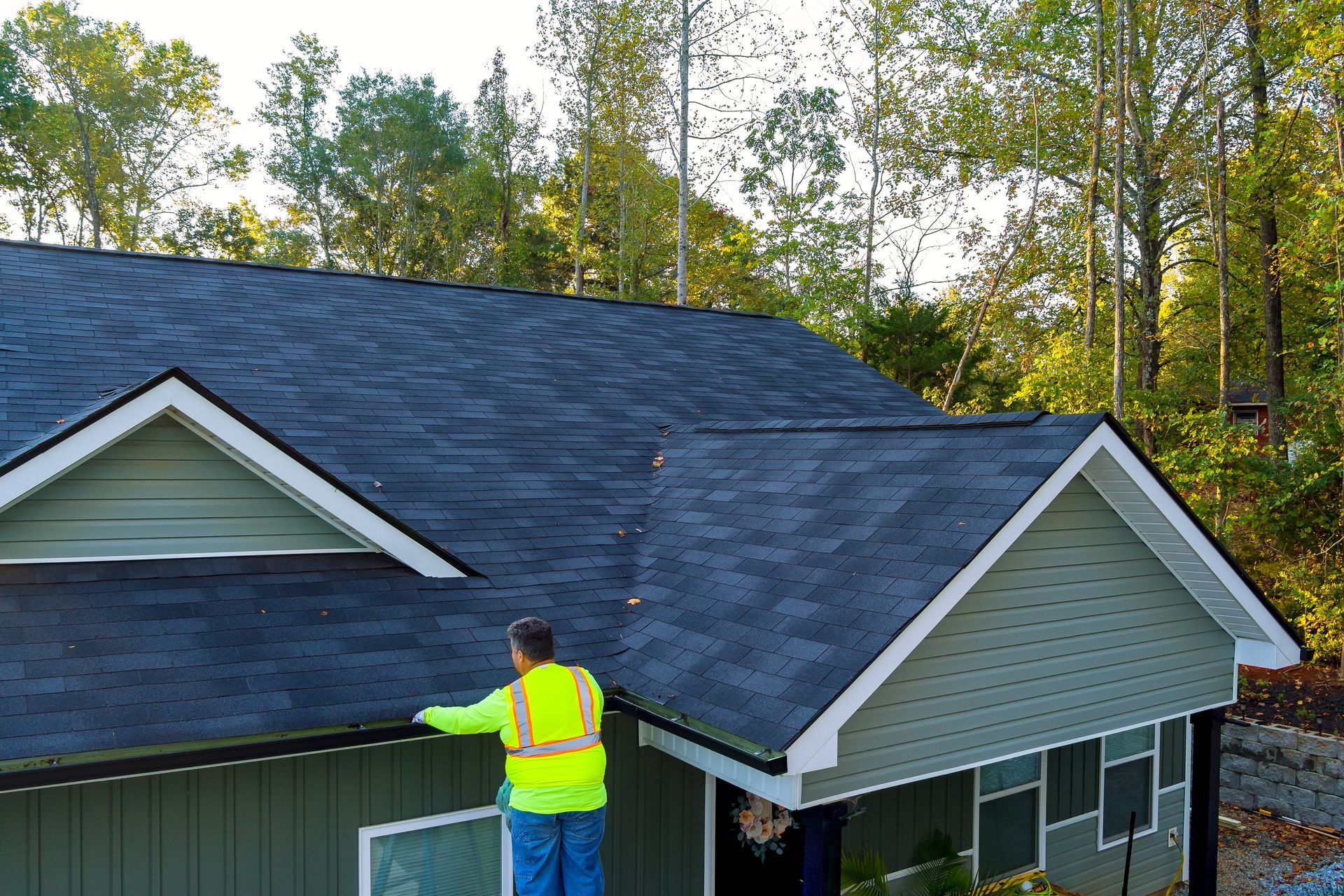 Worker in neon vest inspecting a dark shingled roof on a house surrounded by trees.