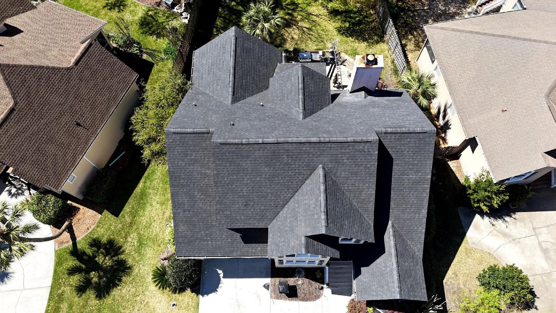 Overhead view of a house with a dark gray shingle roof, surrounded by green trees and other houses.