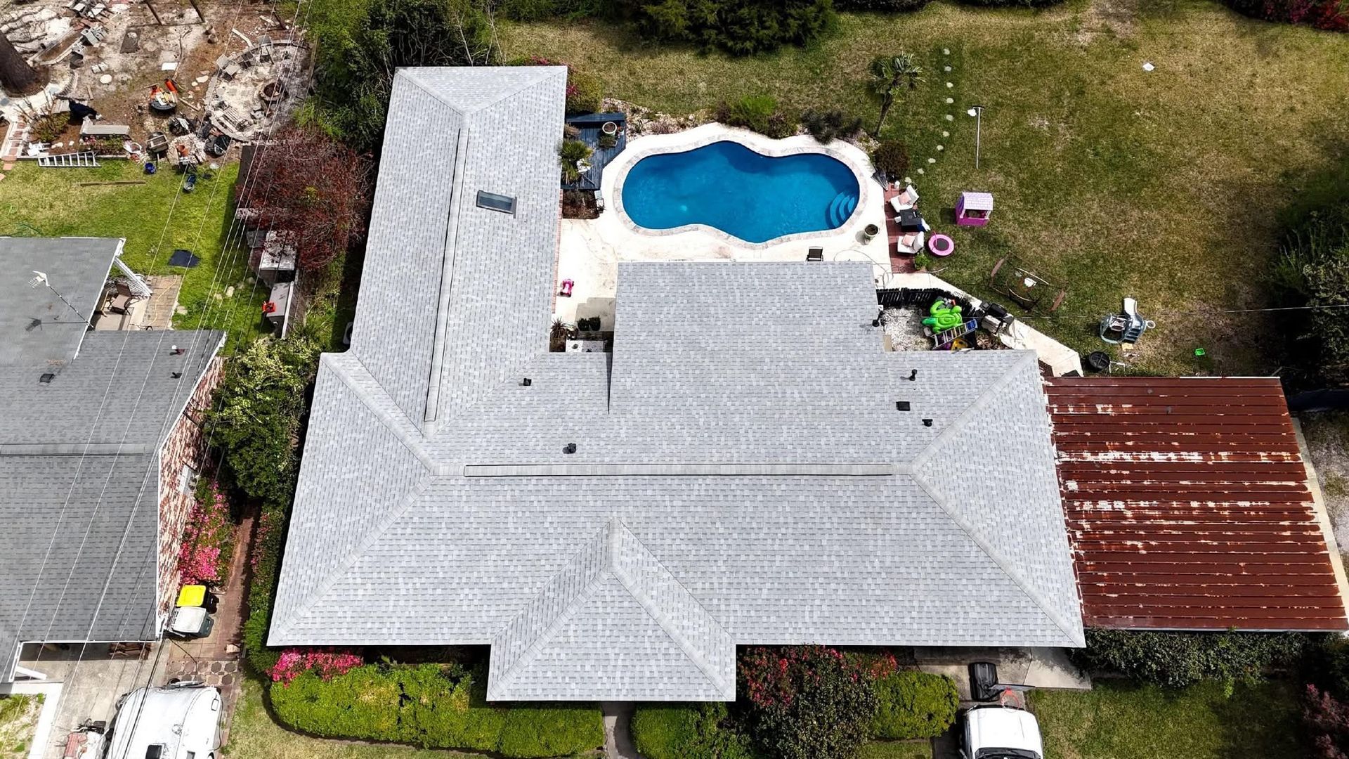 Aerial view of a house with a pool and a corrugated metal roof. Green lawn and trees surround the property.