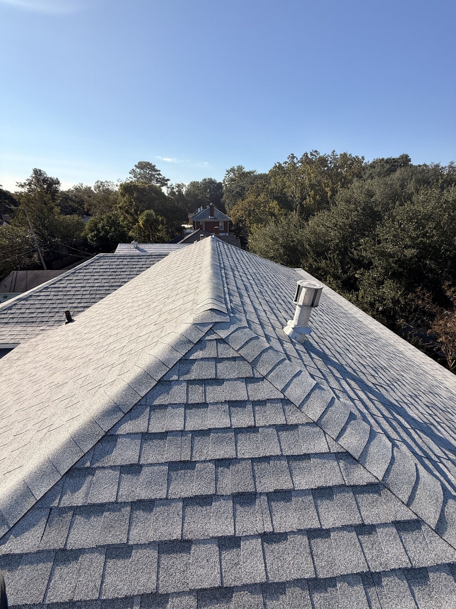 A high-angle view of a gray shingled roof with a chimney, surrounded by green trees under a clear blue sky.