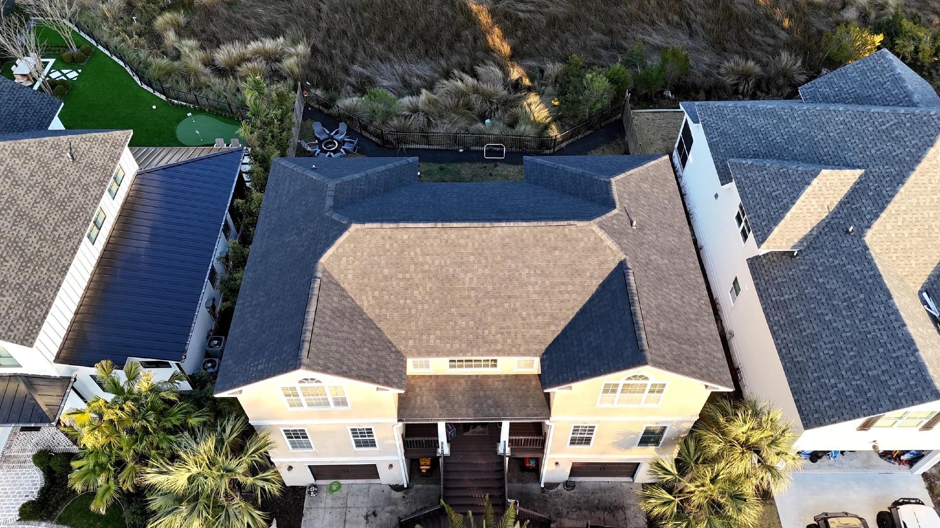 An aerial view of a cream-colored house with a dark brown hip roof, positioned between two adjacent properties.