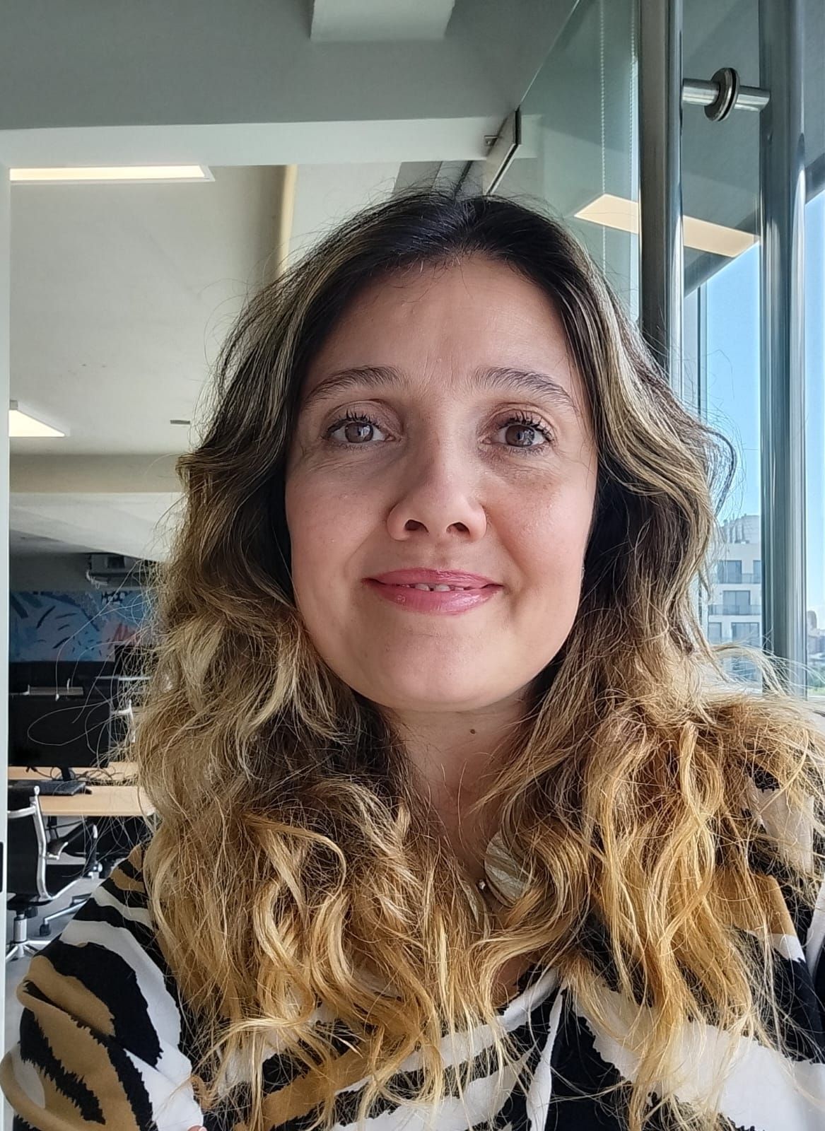 Una mujer sonríe mientras trabaja con una laptop en una mesa redonda blanca junto a una ventana. Hay vasos sobre la mesa.