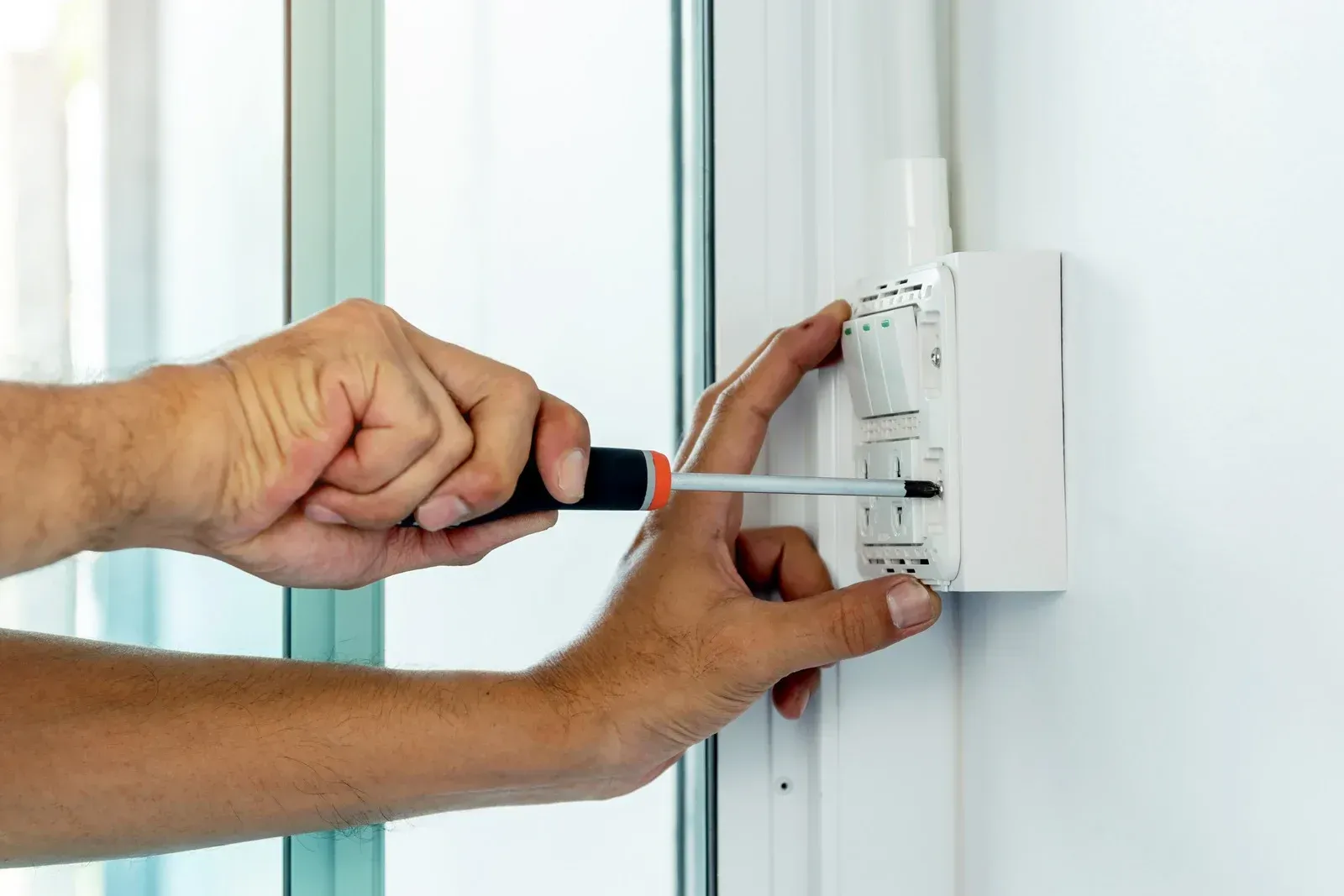 Hands using a screwdriver to install a white light switch on a white wall.