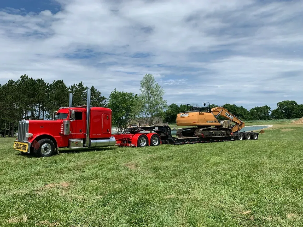 A red semi truck is carrying a bulldozer on a trailer in a grassy field.