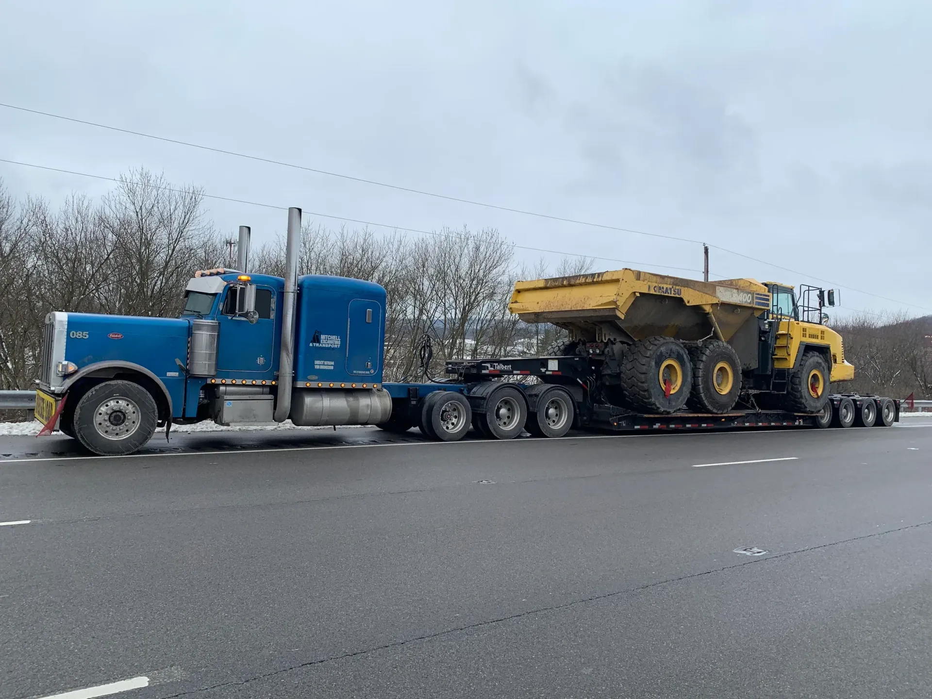 A blue semi truck is carrying a yellow dump truck on a highway.