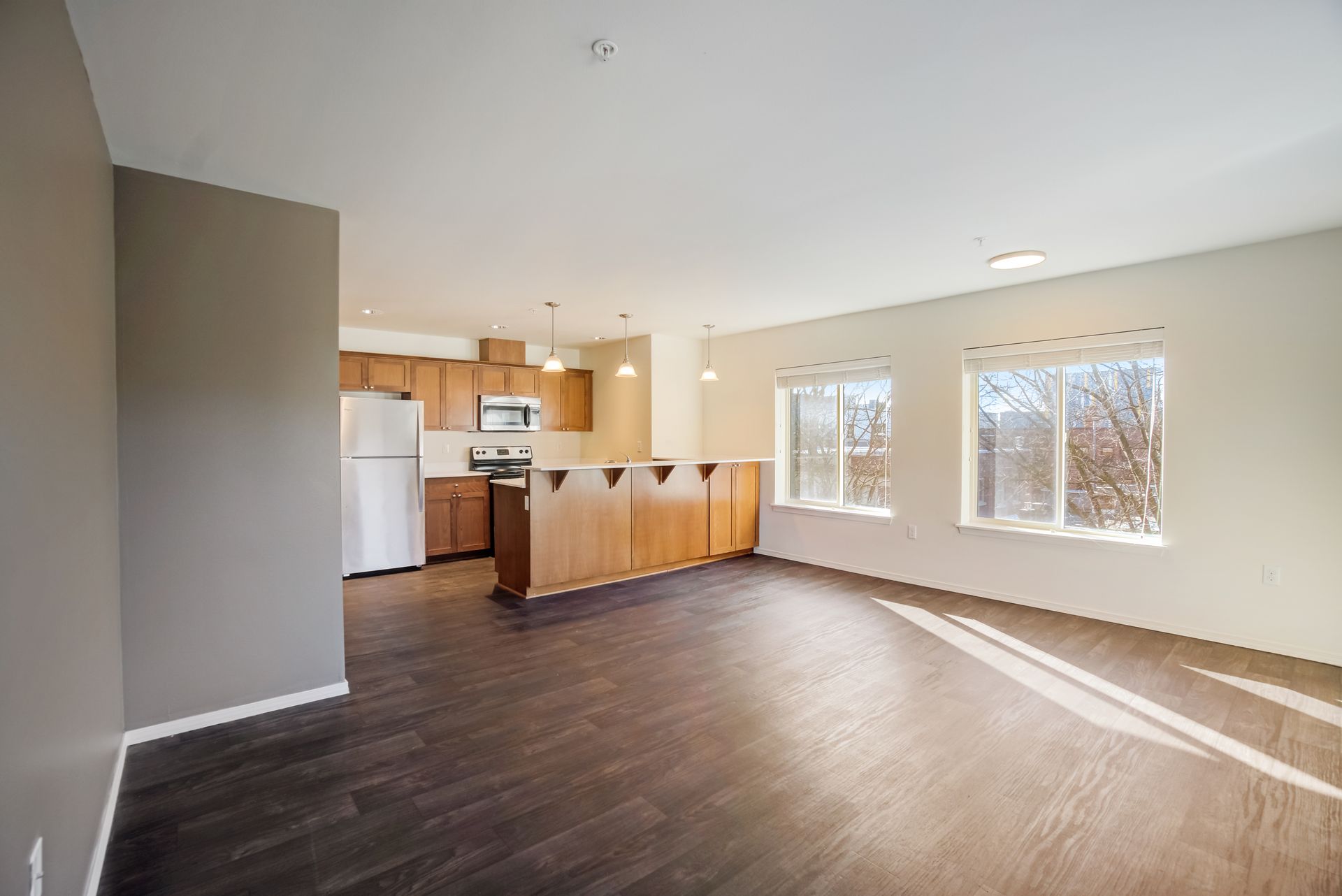 An empty living room with hardwood floors and a kitchen in the background.