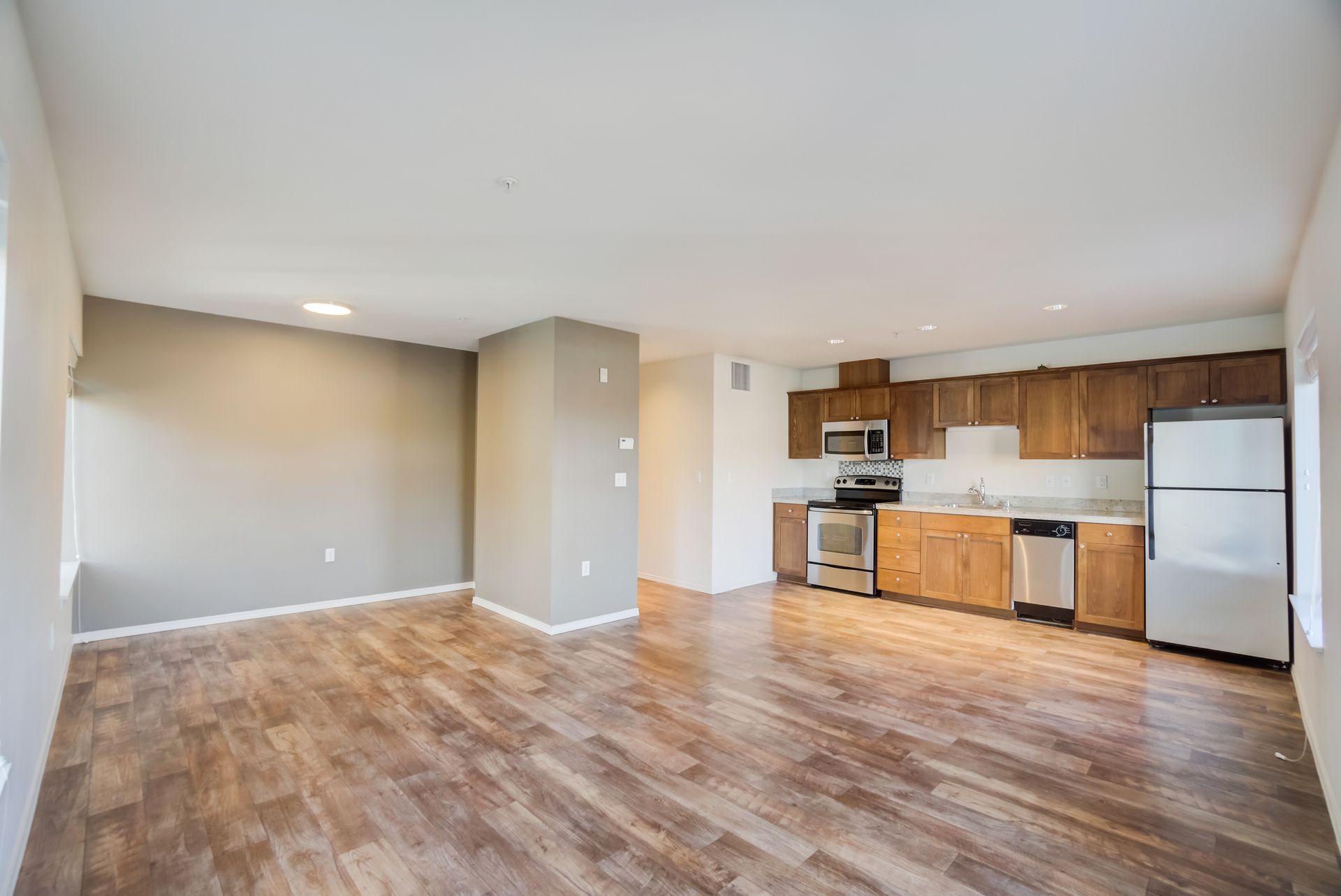 An empty living room with hardwood floors and a kitchen with stainless steel appliances.