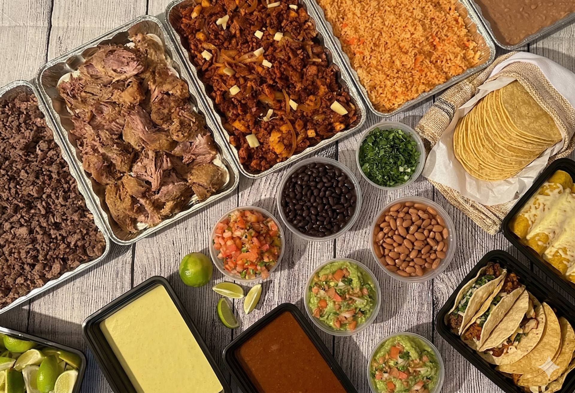 A catering spread featuring a tray of tacos, a tray of taco shells, and a chafing dish of enchiladas on an outdoor table.